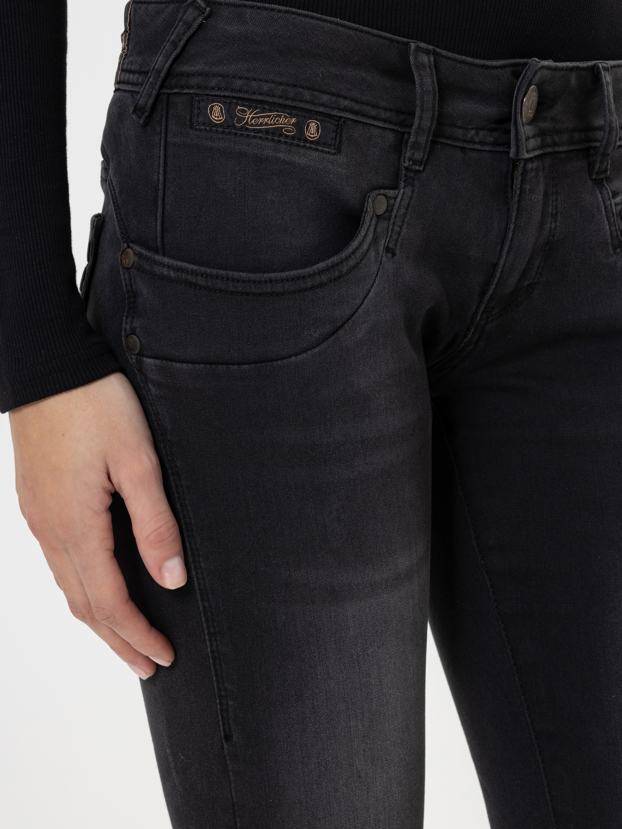 Eine Person, die eine schwarze Jeans und ein schwarzes langärmeliges Oberteil trägt, stützt ihre linke Hand auf den Oberschenkel; die Jeans ist mit Kupfernieten und einem Etikett über der Vordertasche versehen.