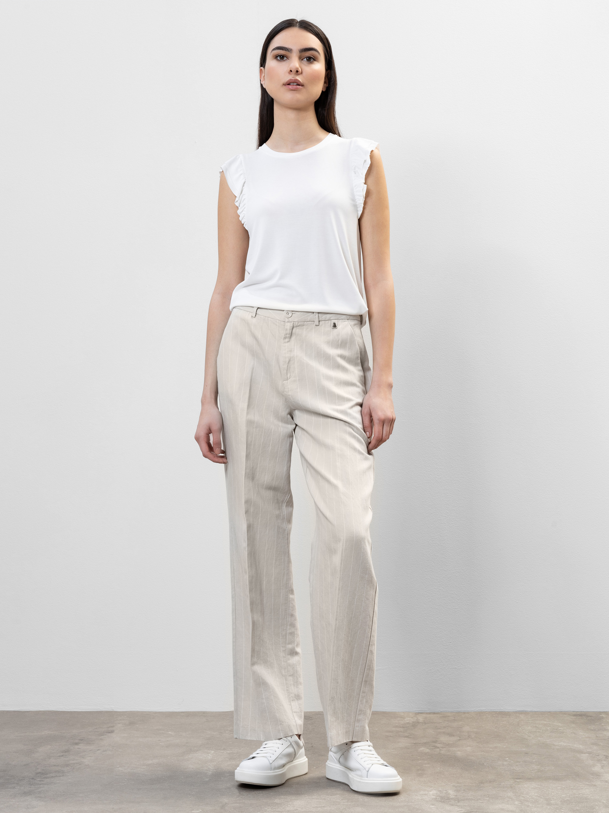 Eine Frau steht vor einem schlichten hellen Hintergrund und trägt ein weißes ärmelloses Rüschen-Top, eine beigefarbene Hose mit geradem Bein und weiße Turnschuhe. Sie hat glattes dunkles Haar und einen neutralen Gesichtsausdruck.