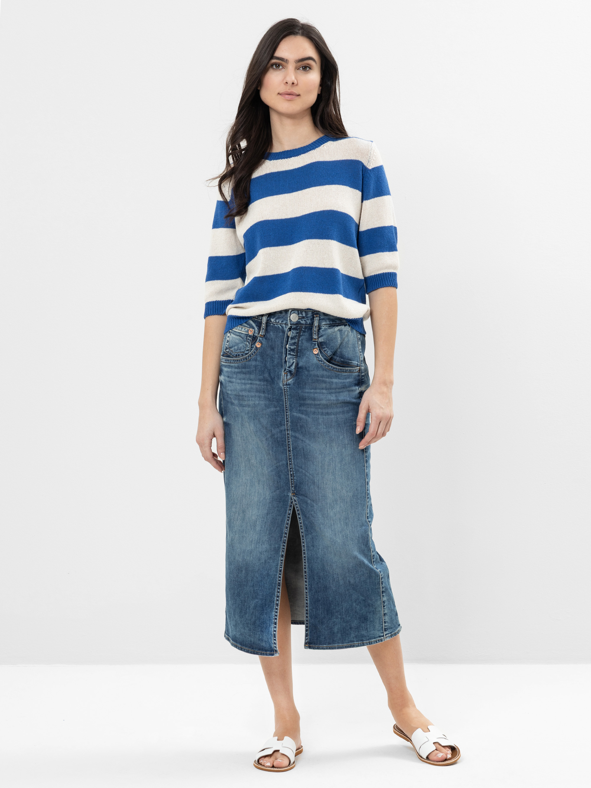 Eine Frau steht vor einem schlichten weißen Hintergrund und trägt einen blau-weiß gestreiften, kurzärmeligen Pullover, einen blauen Jeans-Midirock mit einem Schlitz an der Vorderseite und weiße Slide-Sandalen.