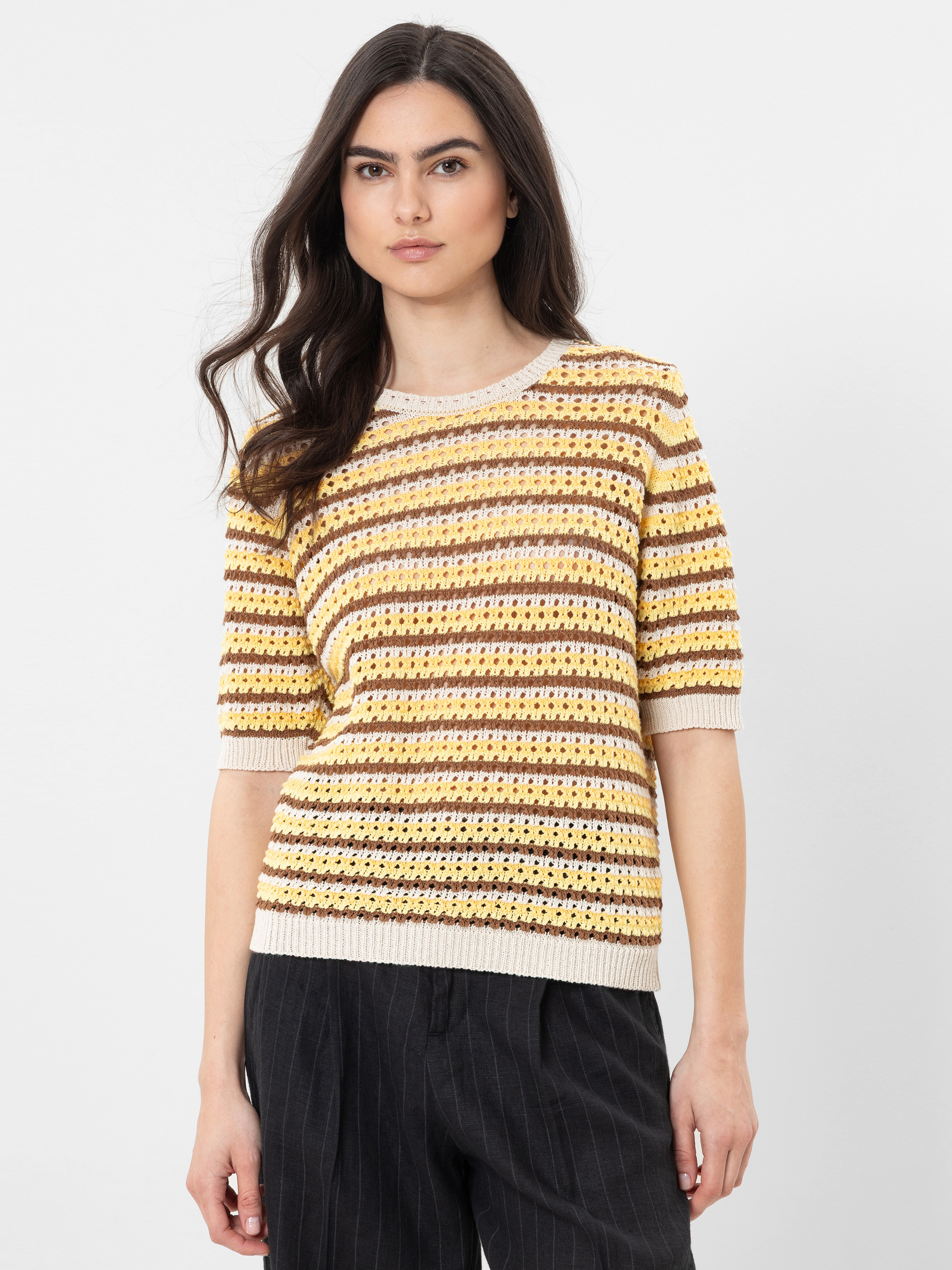 Eine Frau mit langen braunen Haaren trägt einen gelb, braun und weiß gestreiften kurzärmeligen Strickpullover und eine schwarze Nadelstreifenhose und steht vor einem schlichten weißen Hintergrund.