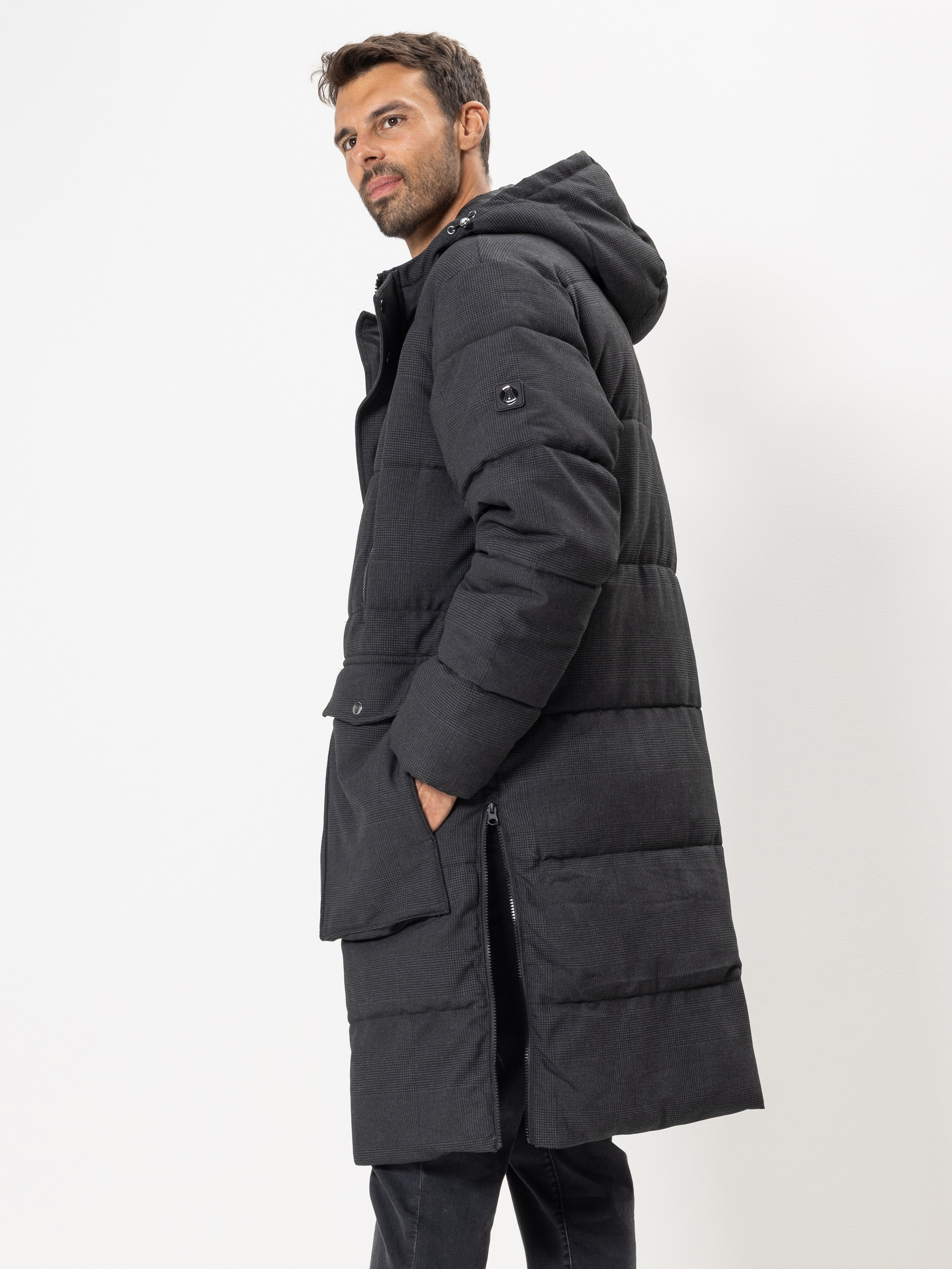 Ein Mann, der einen langen, dunkel gefütterten Wintermantel mit Kapuze trägt, steht seitlich und schaut über seine Schulter. Seine linke Hand steckt in der Manteltasche. Der Hintergrund ist einfarbig weiß.