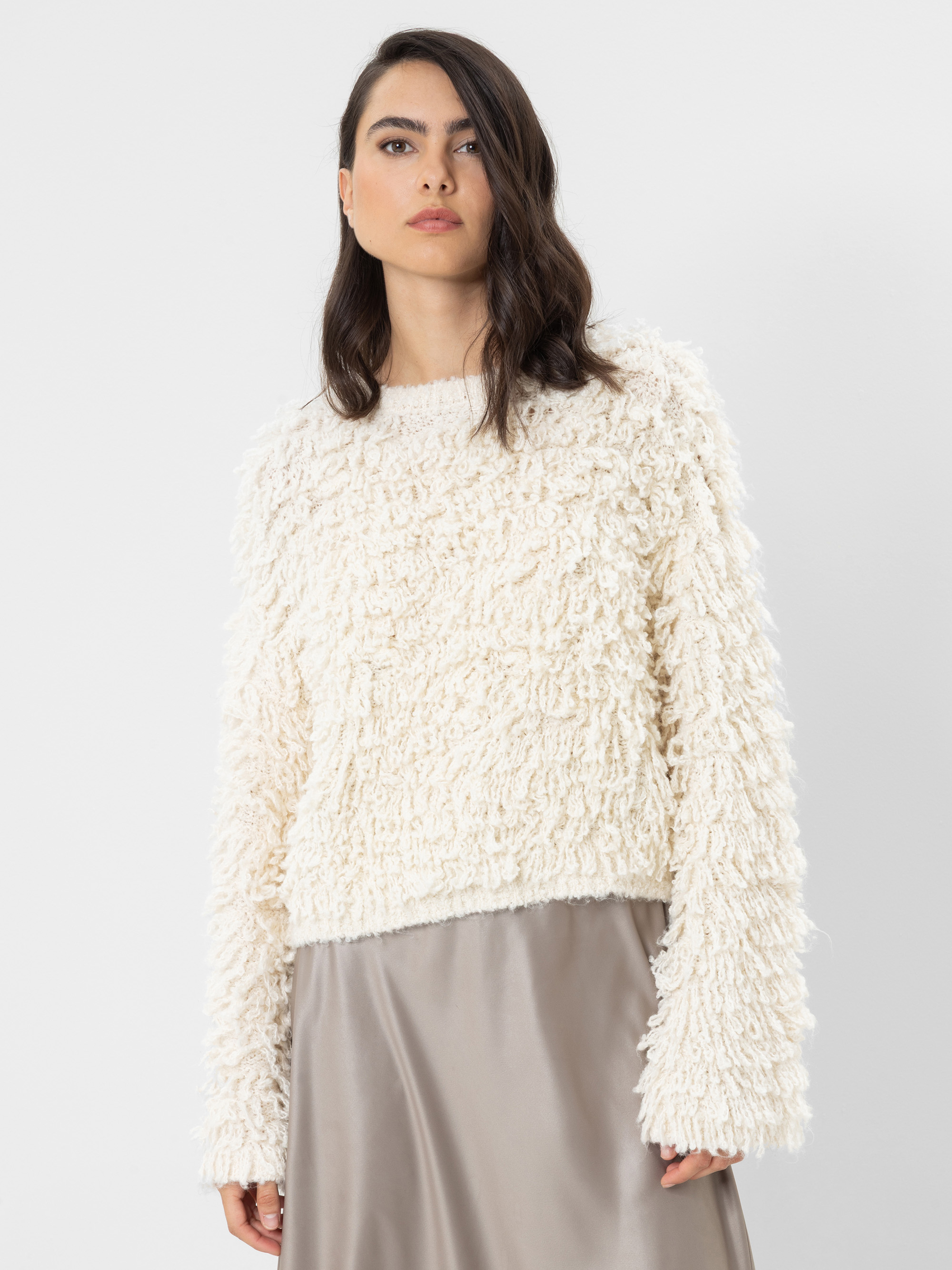 Eine Frau, die einen strukturierten, cremefarbenen, langärmeligen Pullover und einen glänzenden, hellgrauen Rock trägt, steht vor einem schlichten weißen Hintergrund.