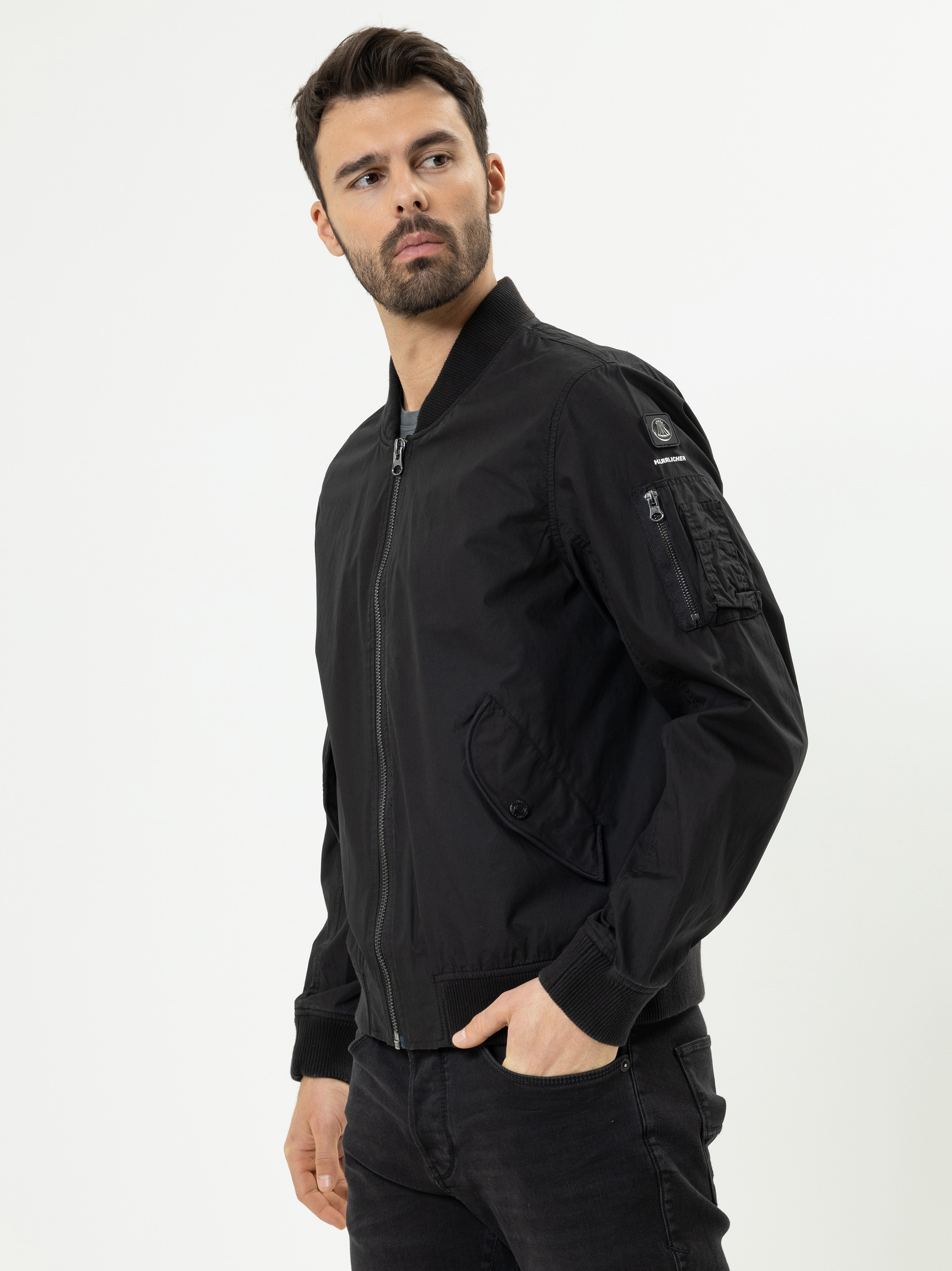 Ein Mann mit kurzen dunklen Haaren und Bart trägt eine schwarze Bomberjacke mit Reißverschluss und schwarze Jeans. Er steht vor einem schlichten weißen Hintergrund und blickt leicht nach links, eine Hand in der Tasche.