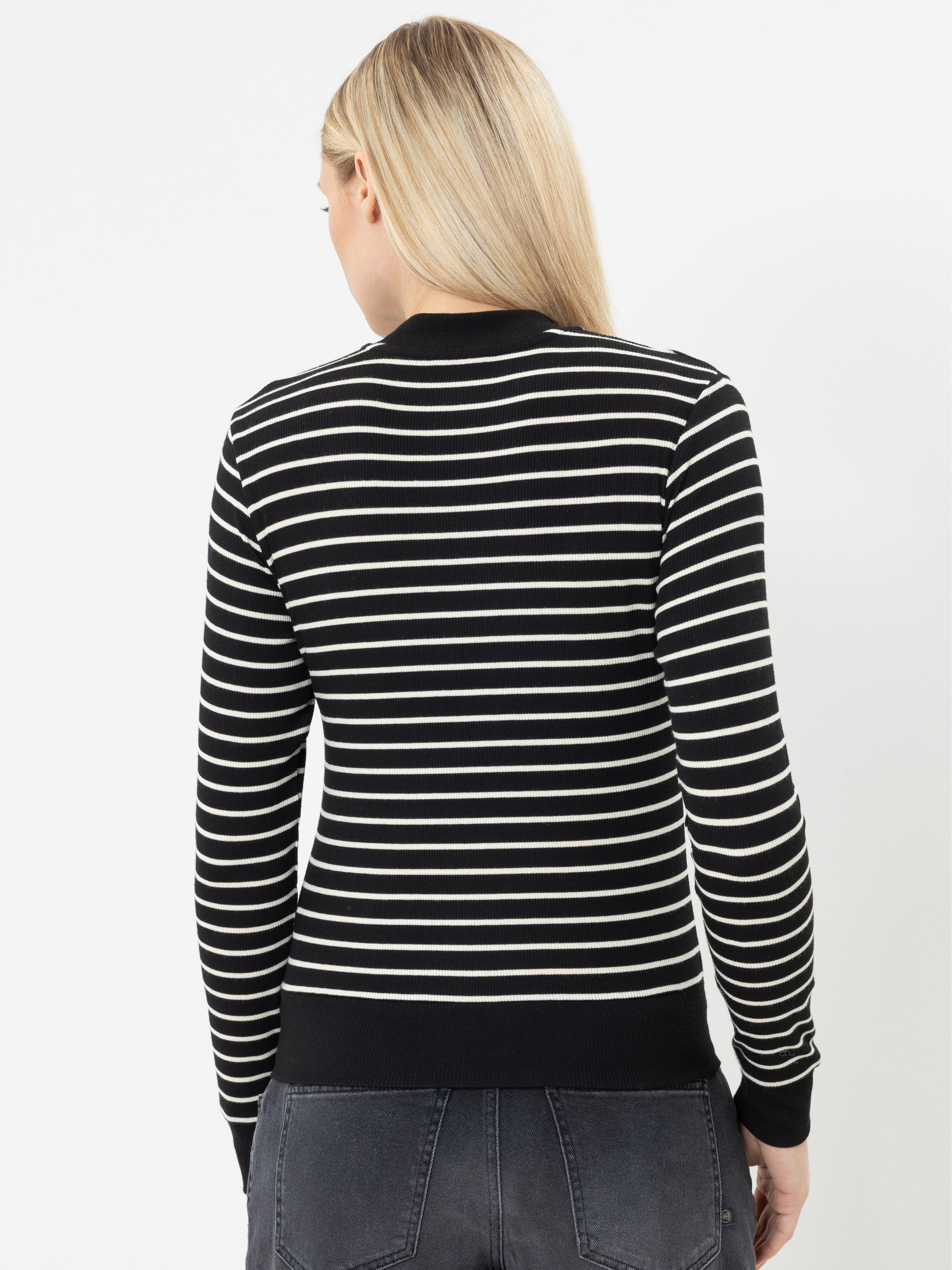 Eine Frau mit langen blonden Haaren steht mit dem Rücken zur Kamera, trägt einen schwarzen Pullover mit weißen Querstreifen und eine dunkle Jeans und steht vor einem schlichten weißen Hintergrund.