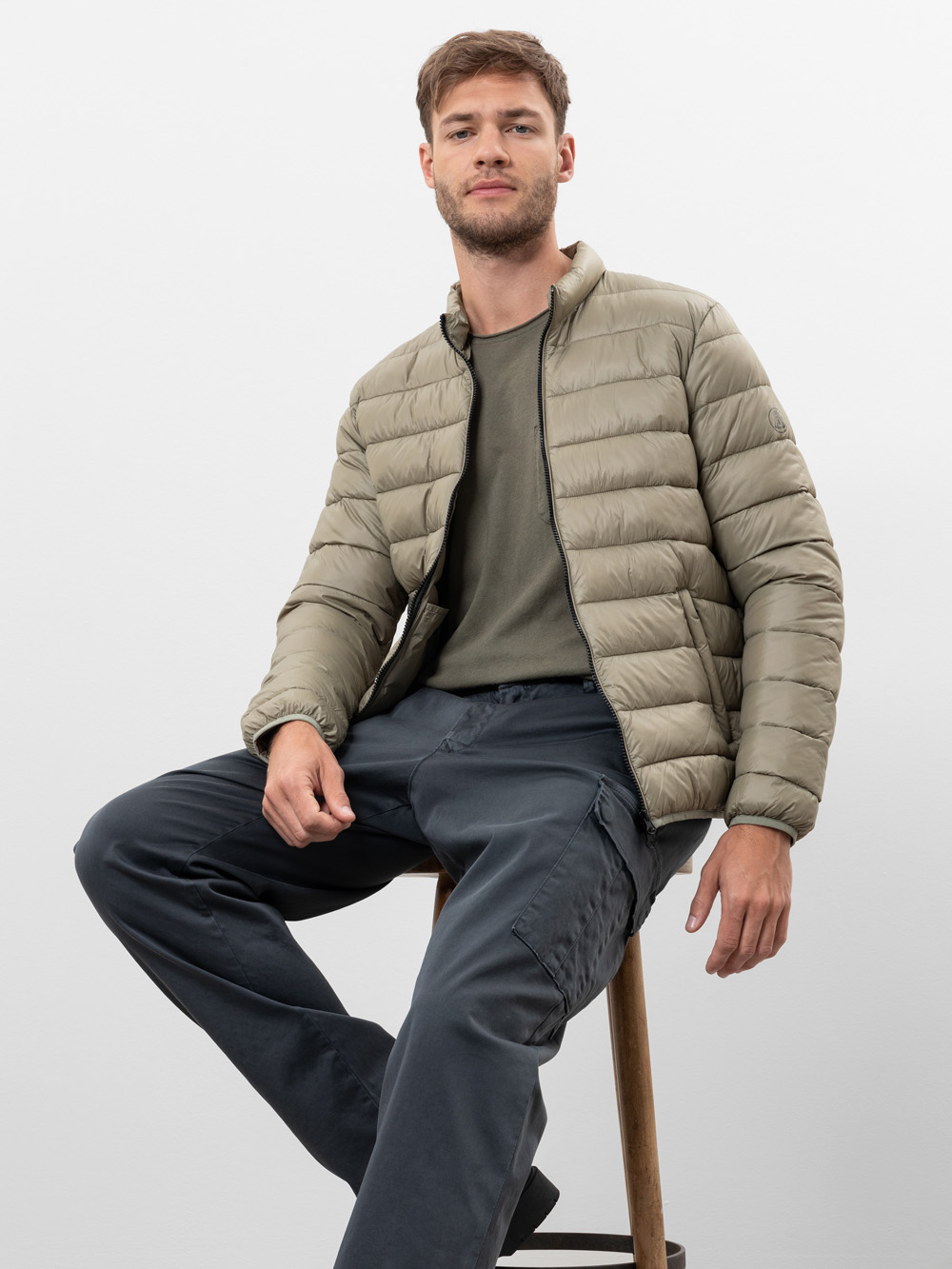 Ein Mann mit kurzen braunen Haaren sitzt auf einem Holzschemel und trägt eine hellolivfarbene Pufferjacke über einem olivgrünen T-Shirt und eine dunkelblaue Cargohose vor einem schlichten weißen Hintergrund.