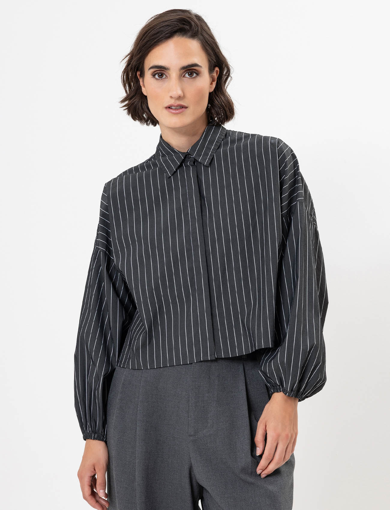Eine Person, die ein lockeres, dunkelgraues, langärmeliges Hemd mit Kragen und vertikalen weißen Nadelstreifen und eine dazu passende graue Hose mit hoher Taille trägt, steht vor einem schlichten weißen Hintergrund.