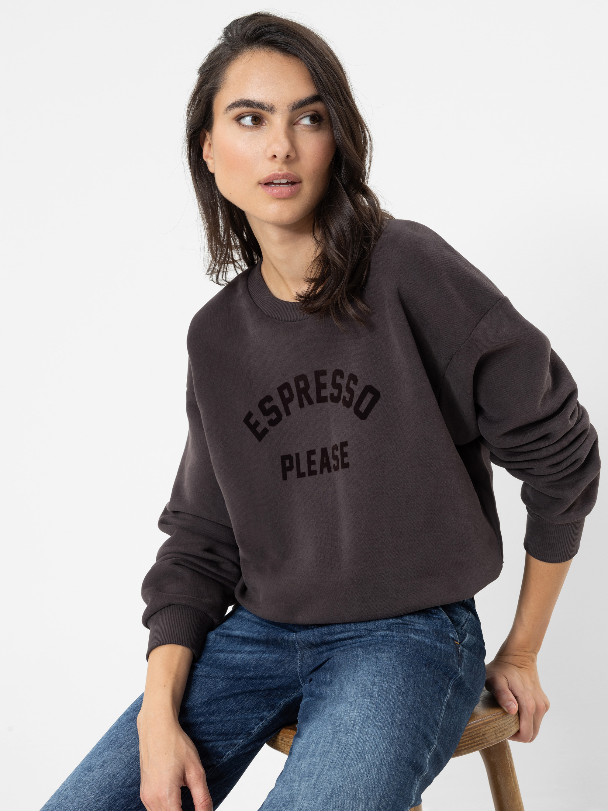 Vor einem schlichten weißen Hintergrund sitzt eine Frau mit langen braunen Haaren auf einem Holzhocker; sie trägt ein dunkles Sweatshirt mit der Aufschrift „ESPRESSO PLEASE“ auf der Vorderseite und eine blaue Jeans.