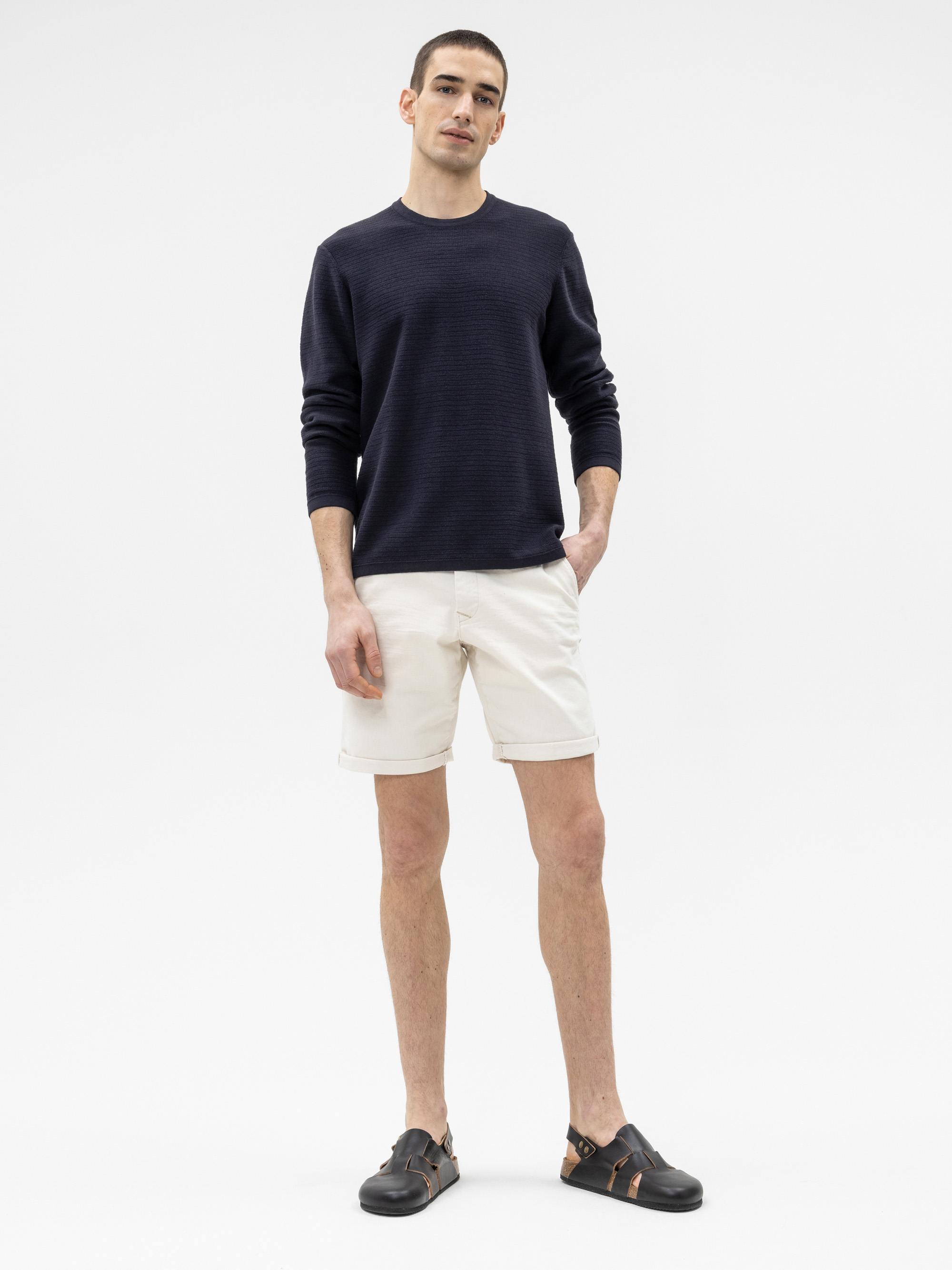 Ein Mann steht vor einem weißen Hintergrund und trägt ein dunkles langärmeliges Hemd, helle Shorts und schwarze Sandalen. Er hat kurze Haare und seine linke Hand steckt in der Tasche.