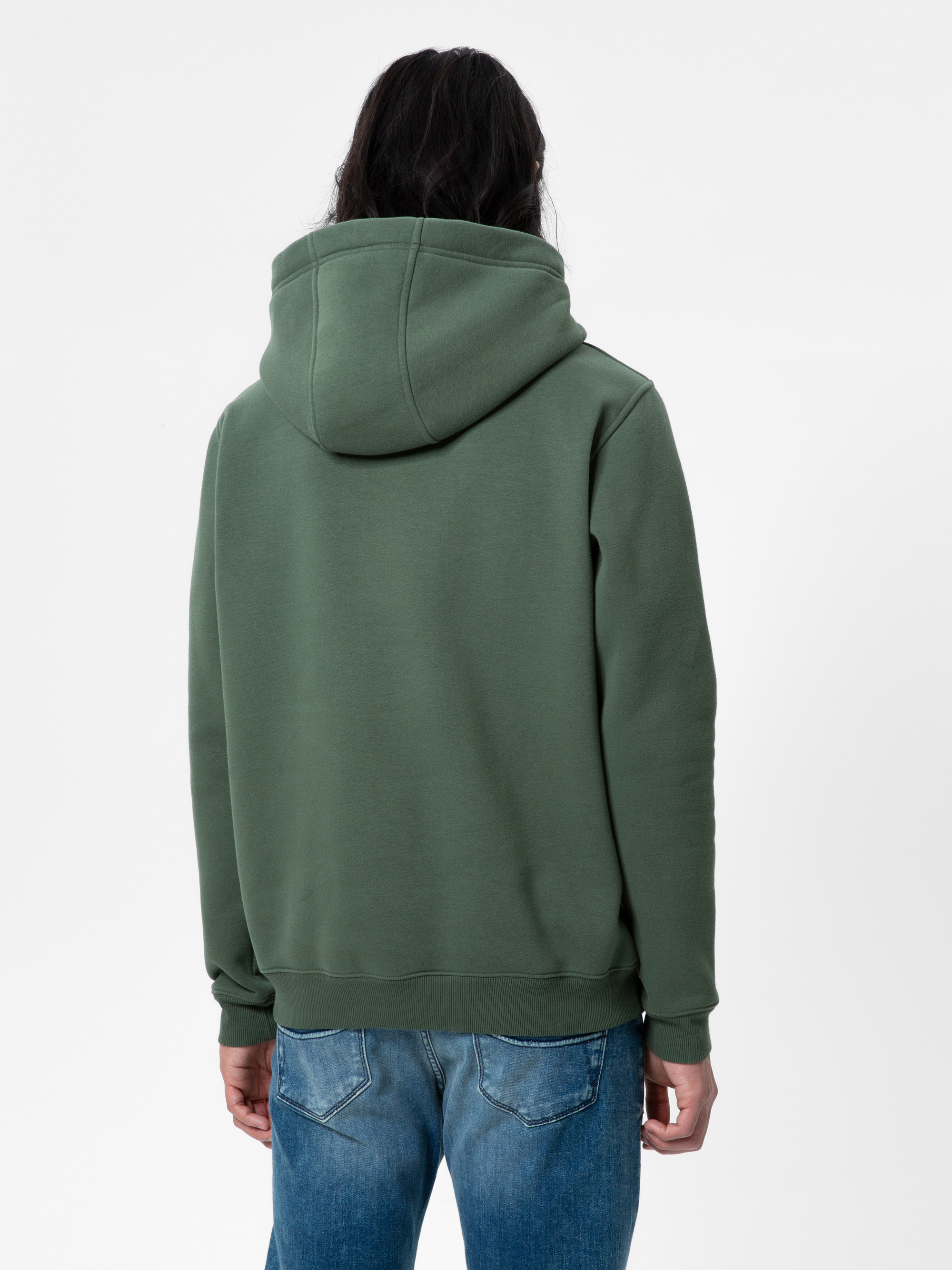 Eine Person mit langen dunklen Haaren steht mit dem Gesicht nach hinten, trägt ein grünes Kapuzensweatshirt und blaue Jeans vor einem schlichten weißen Hintergrund.