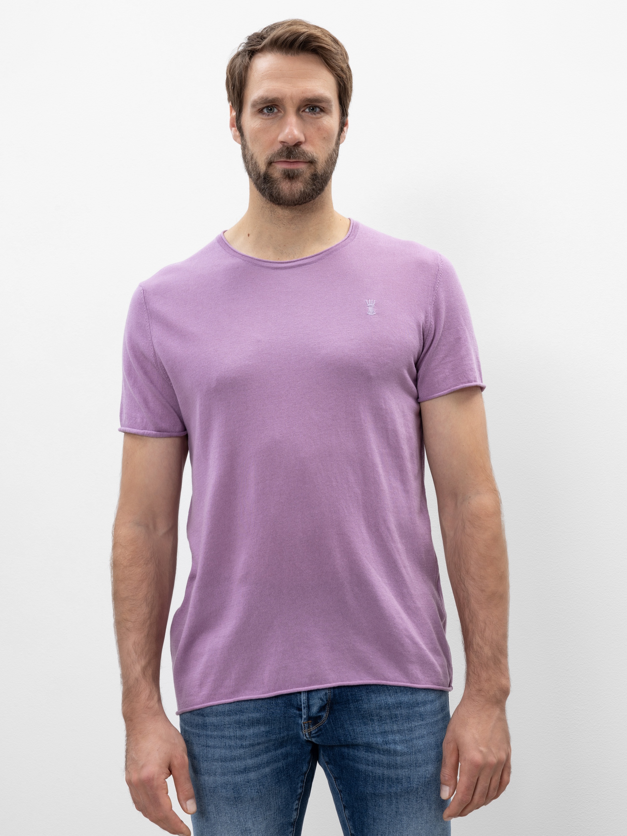 Ein Mann mit hellbraunem Haar und Bart steht vor einem schlichten weißen Hintergrund und trägt ein hellviolettes T-Shirt und blaue Jeans. Er blickt nach vorne und hat einen neutralen Gesichtsausdruck.