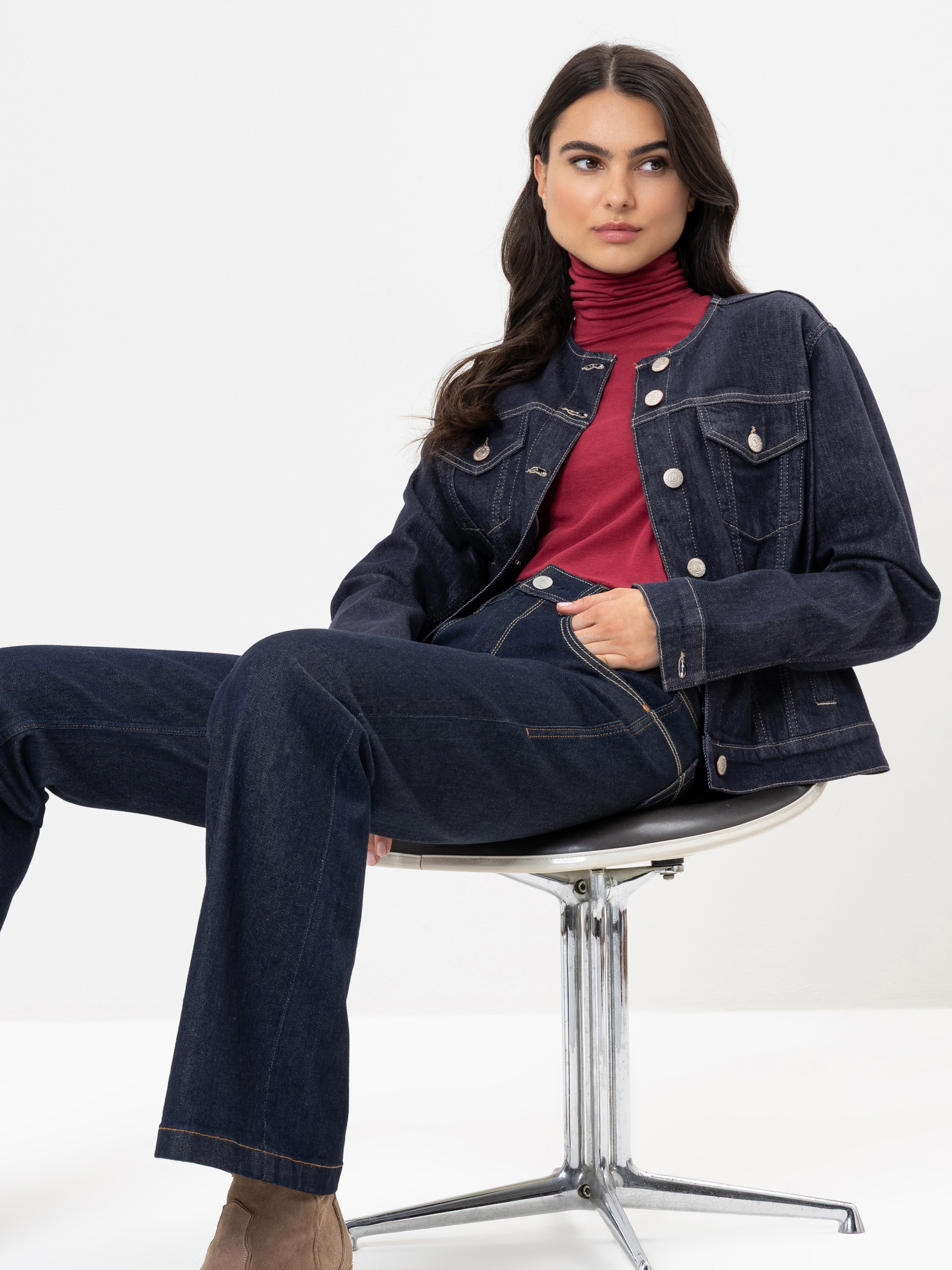 Eine Frau mit langen dunklen Haaren sitzt auf einem modernen Stuhl. Sie trägt eine dunkle Jeansjacke und Jeans mit einem roten Rollkragenpullover. Sie blickt selbstbewusst in die Kamera und hat eine Hand in der Tasche.