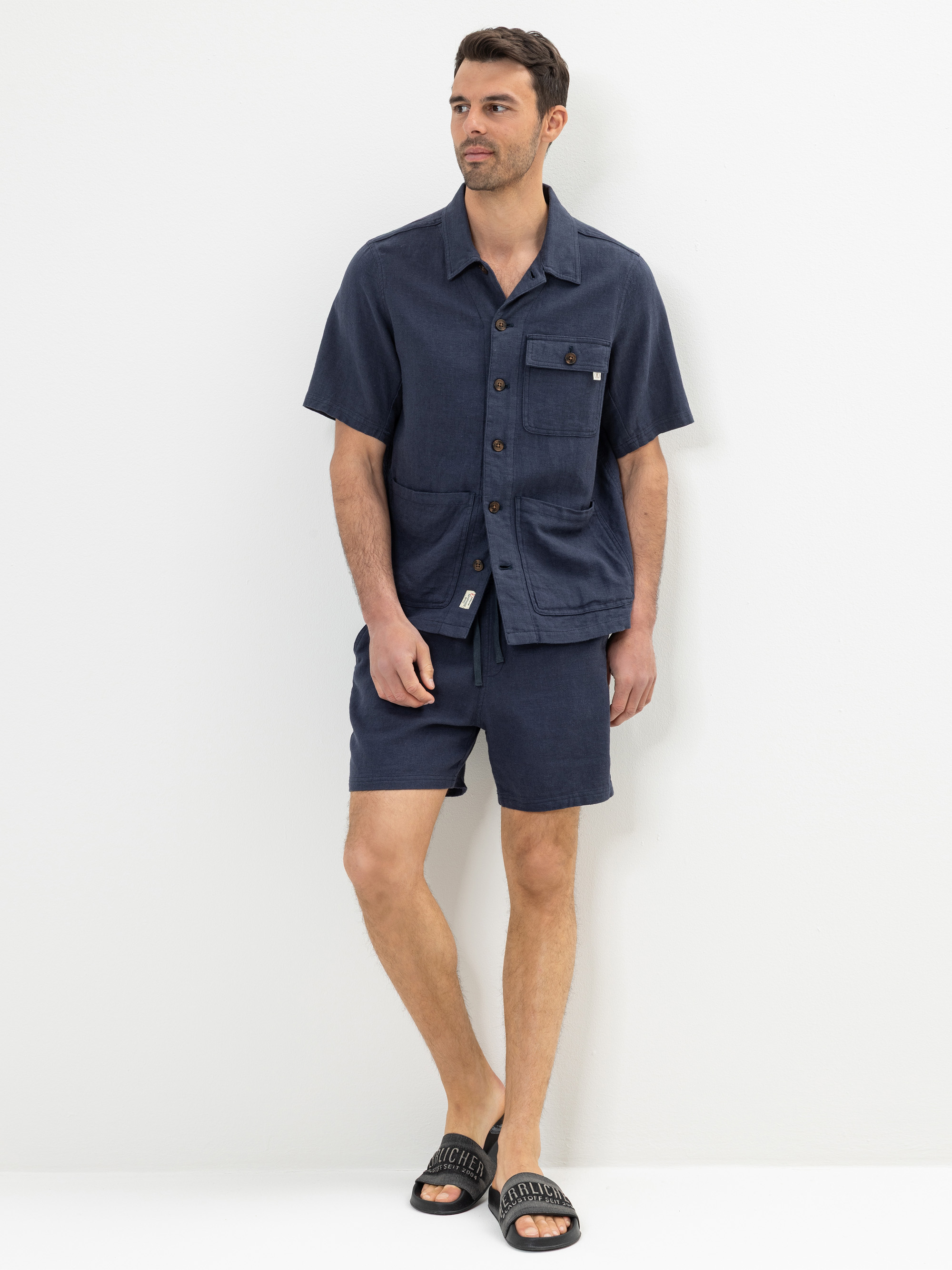 Ein Mann steht vor einer weißen Wand und trägt ein marineblaues, kurzärmeliges Button-up-Hemd mit Vordertaschen, passende marineblaue Shorts und schwarze Slide-Sandalen. Er blickt leicht zur Seite und hat einen entspannten Gesichtsausdruck.