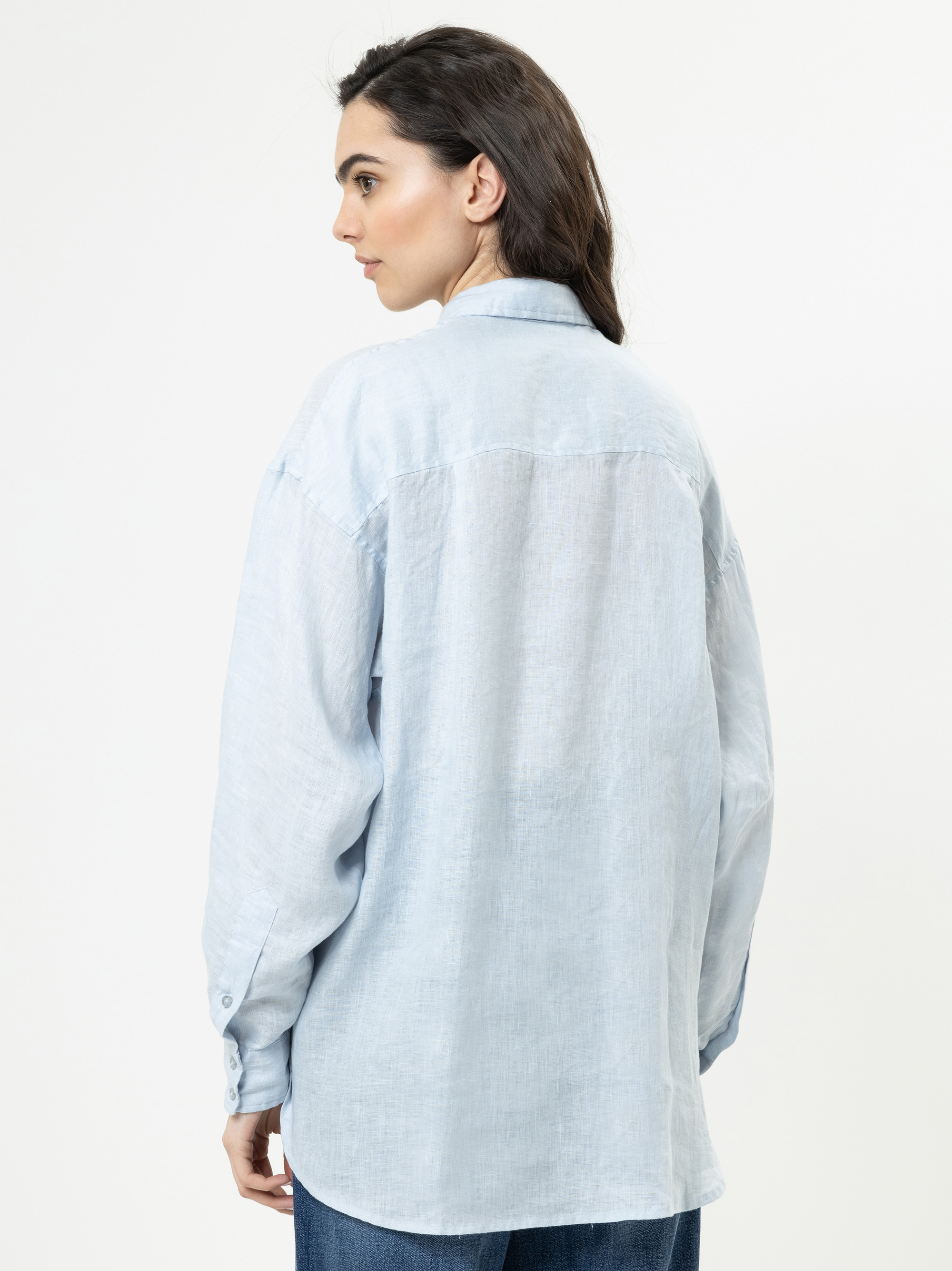 Eine Frau mit langen dunklen Haaren, die ein hellblaues, locker sitzendes Button-up-Hemd trägt, steht mit dem Gesicht zur Seite und blickt vor einem schlichten weißen Hintergrund.