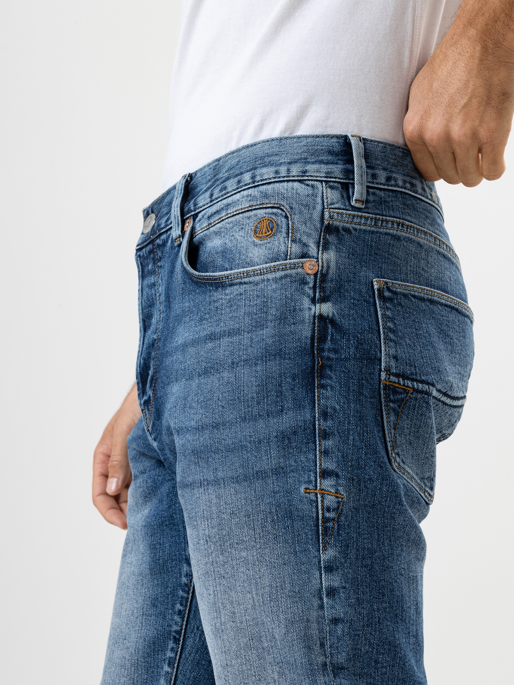 Eine Person, die eine blaue Jeans und ein weißes T-Shirt trägt, wird von der Taille abwärts gezeigt, wobei eine Hand den Bund der Jeans vor einem schlichten hellen Hintergrund zurechtrückt.