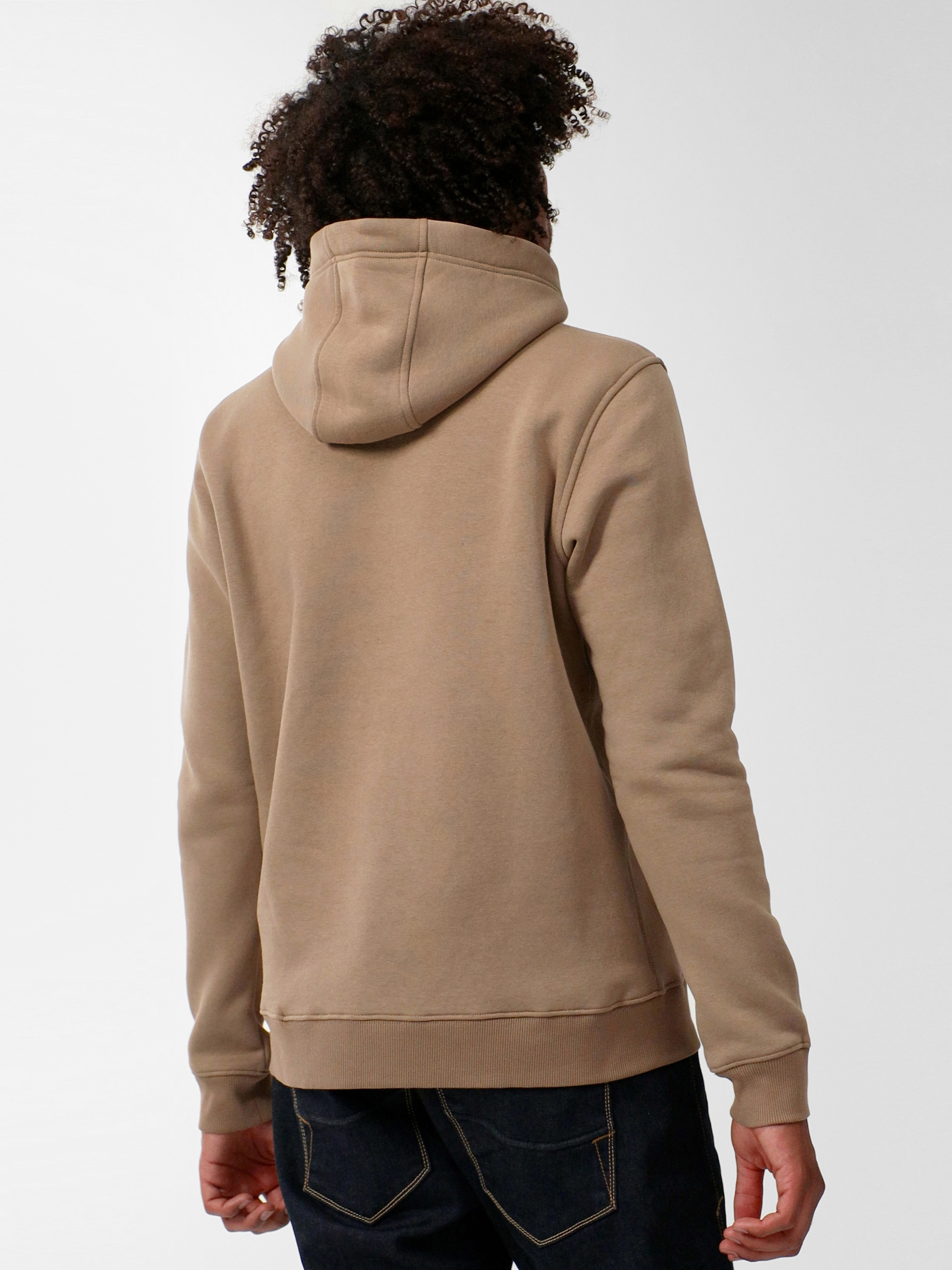 Eine Person mit lockigem Haar steht mit dem Rücken zur Kamera, trägt einen hellbraunen Kapuzenpullover und dunkelblaue Jeans vor einem schlichten, hellen Hintergrund.