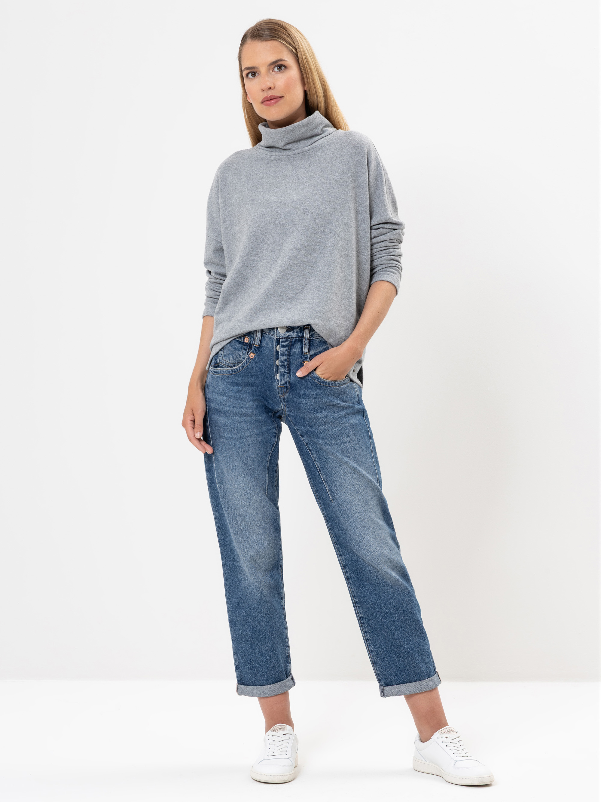 Eine Frau steht vor einem weißen Hintergrund und trägt einen hellgrauen Rollkragenpullover, blaue Jeans mit Bündchen und weiße Turnschuhe. Sie hat glattes, langes Haar und blickt leicht zur Seite, eine Hand in der Tasche.