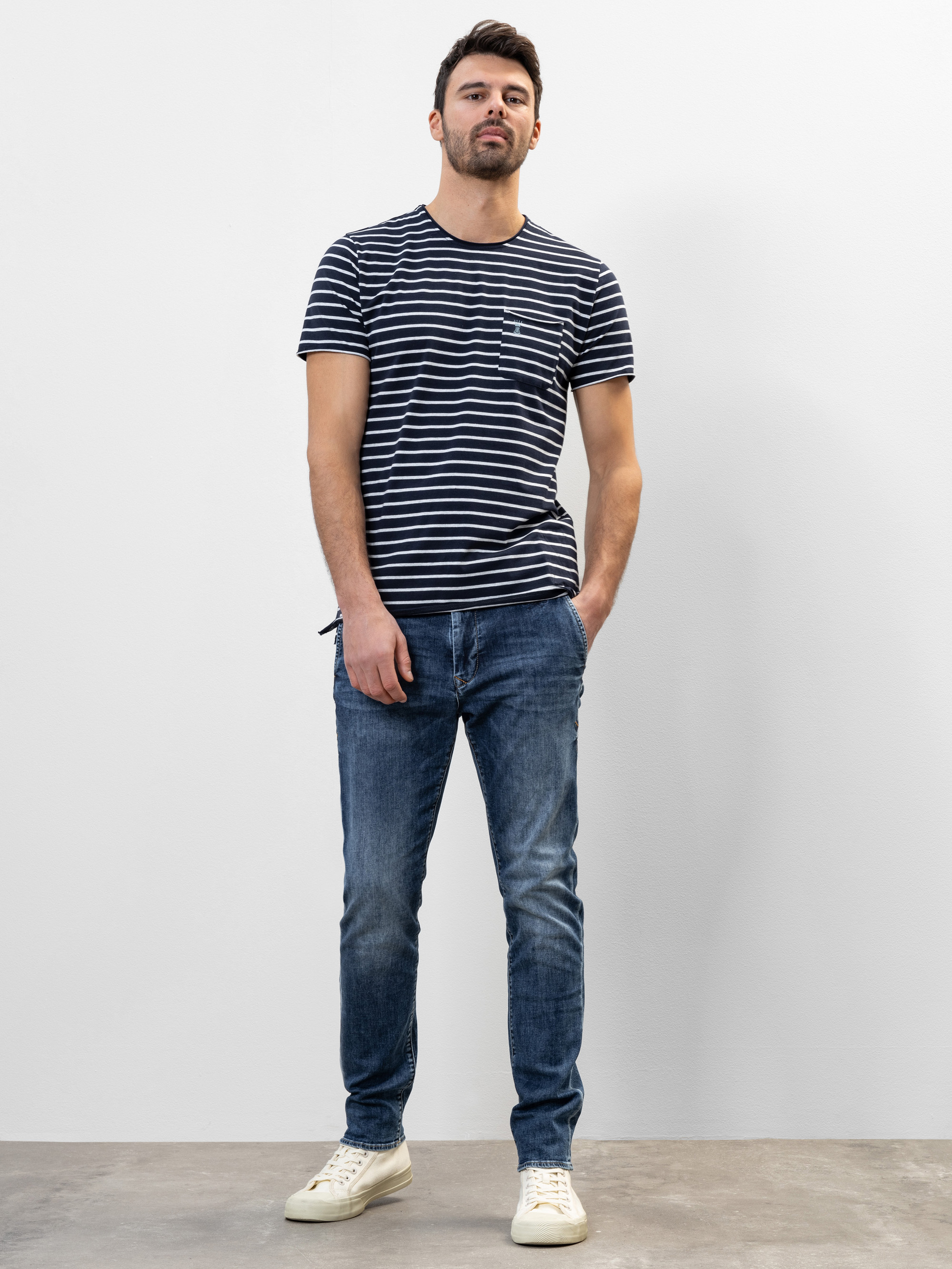 Ein Mann steht vor einem schlichten weißen Hintergrund und trägt ein marineblau-weiß gestreiftes T-Shirt, blaue Jeans und weiße Turnschuhe. Eine Hand steckt in seiner Tasche und er hat einen neutralen Gesichtsausdruck.