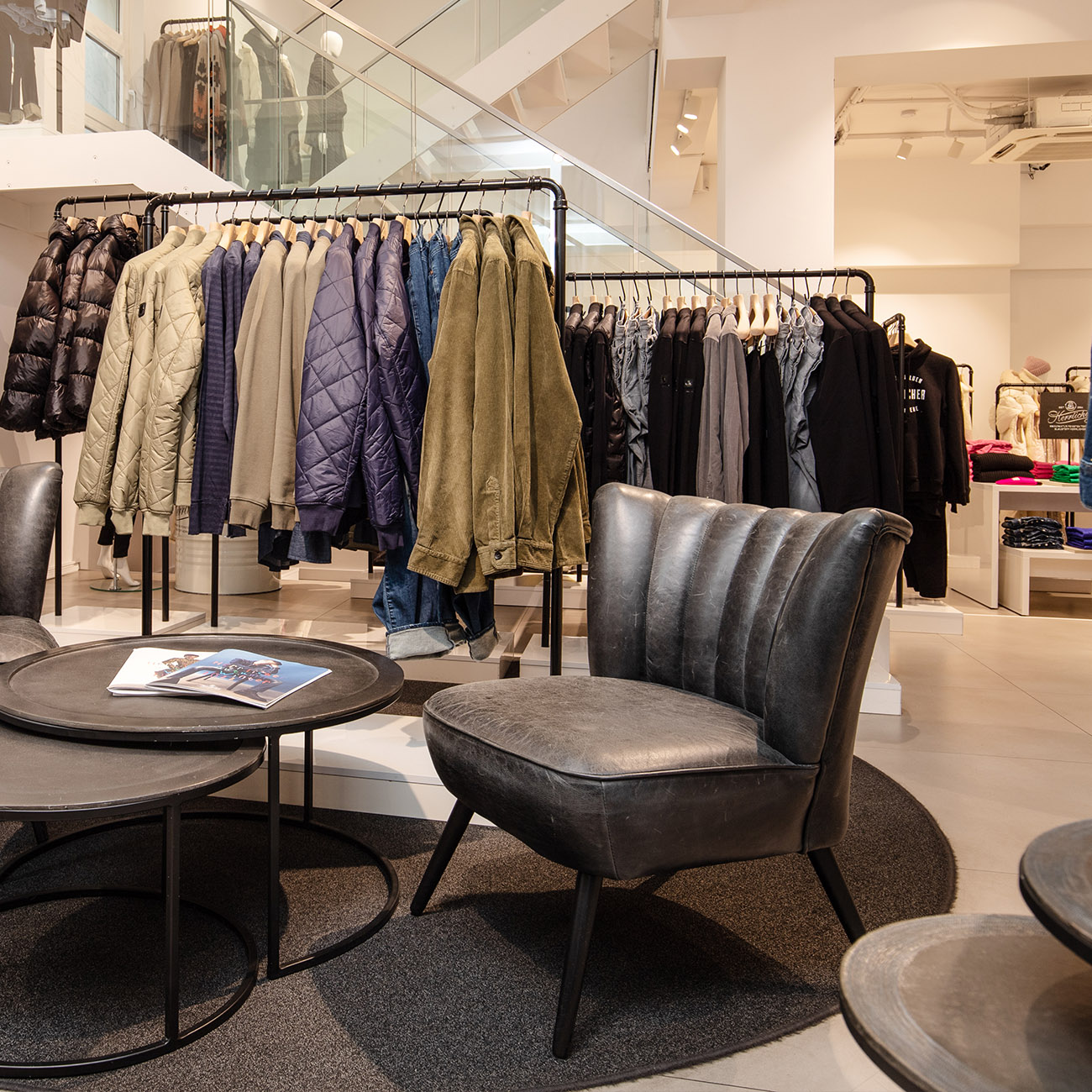 Ein modernes Bekleidungsgeschäft mit Regalen mit Jacken und Hemden, einem runden Tisch mit einer Zeitschrift und einem grauen gepolsterten Stuhl auf einem runden Teppich in der Nähe einer Treppe mit Glasgeländer.