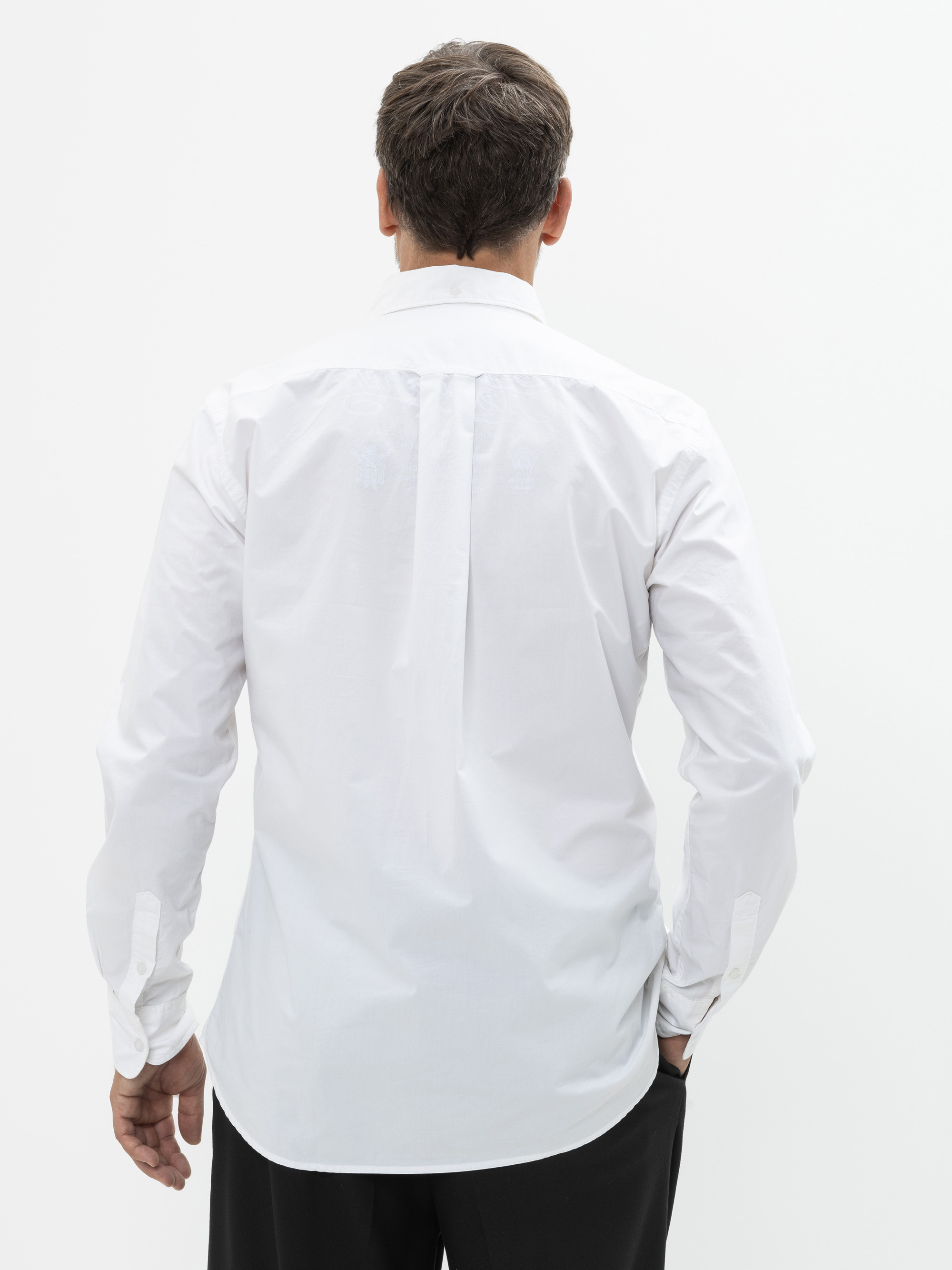 Ein Mann mit kurzen braunen Haaren steht mit dem Rücken zur Kamera. Er trägt ein weißes langärmeliges Button-up-Hemd und eine schwarze Hose vor einem schlichten weißen Hintergrund.