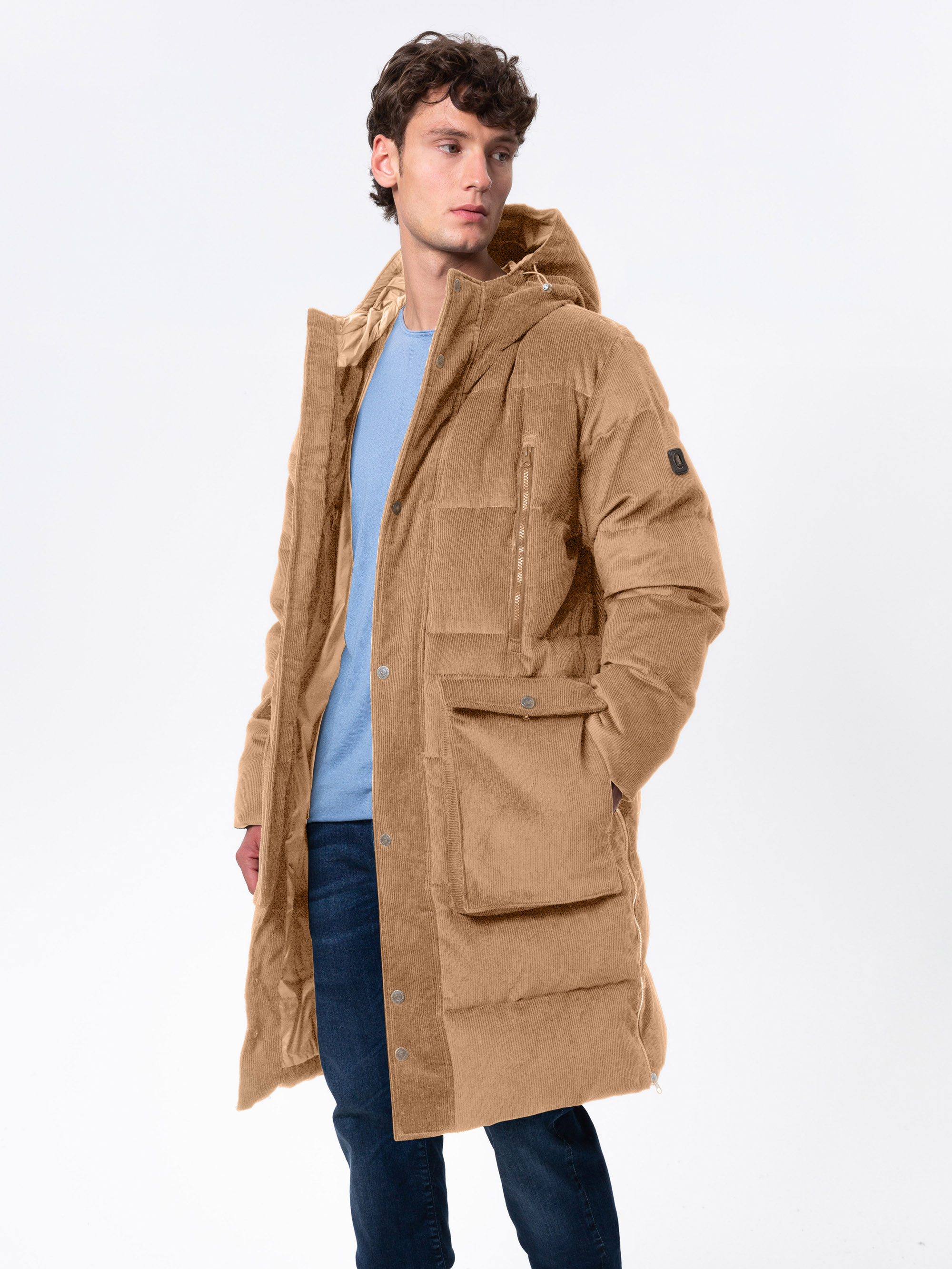 Ein Mann, der einen hellbraunen, knielangen Wintermantel mit Kapuze über einem hellblauen Pullover und dunkle Jeans trägt, steht vor einem schlichten weißen Hintergrund und blickt zur Seite.