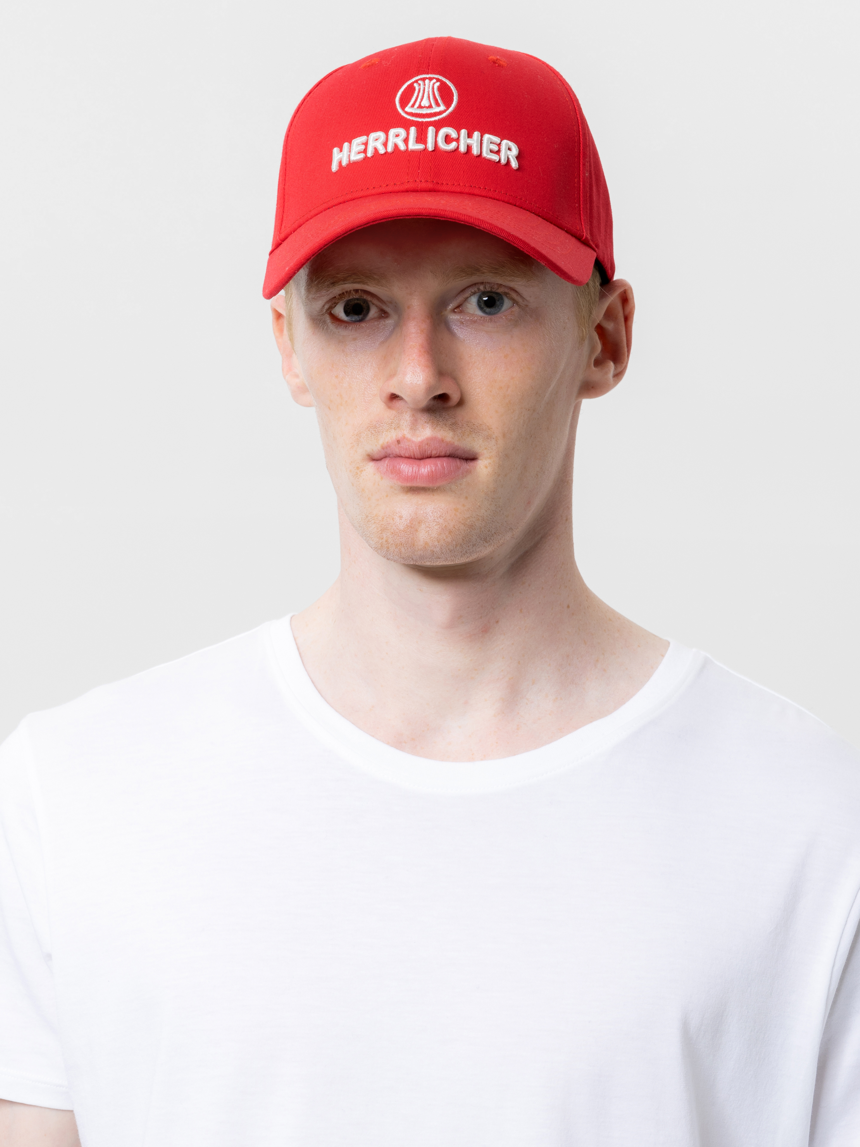 Eine Person, die ein einfaches weißes T-Shirt und eine rote Mütze mit dem Aufdruck HERRLICHER trägt, steht vor einem einfachen weißen Hintergrund.