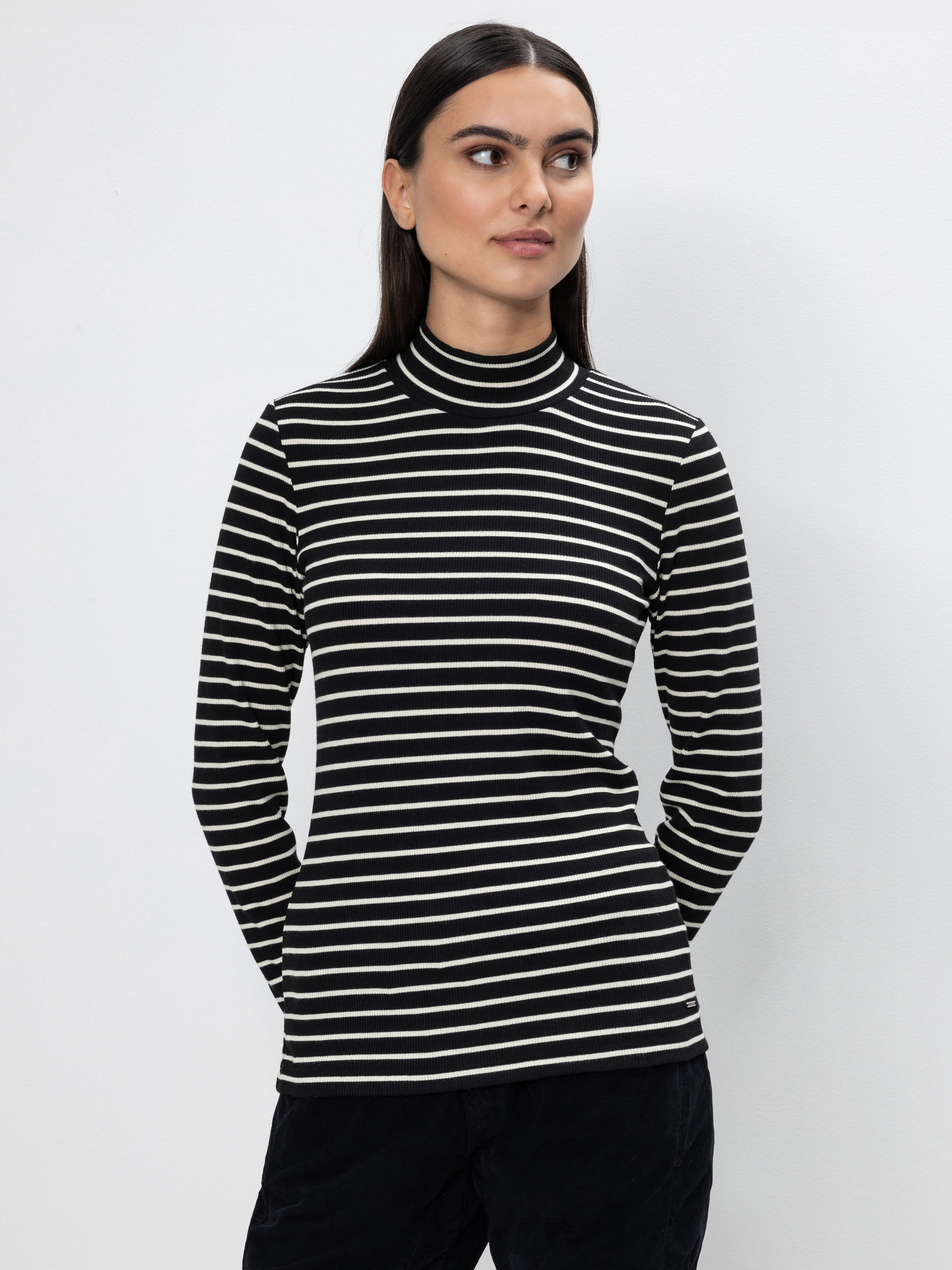 Eine Frau mit glattem dunklem Haar trägt einen schwarz-weiß horizontal gestreiften Rollkragenpullover und eine schwarze Hose und steht mit den Händen in den Taschen vor einem schlichten hellen Hintergrund.