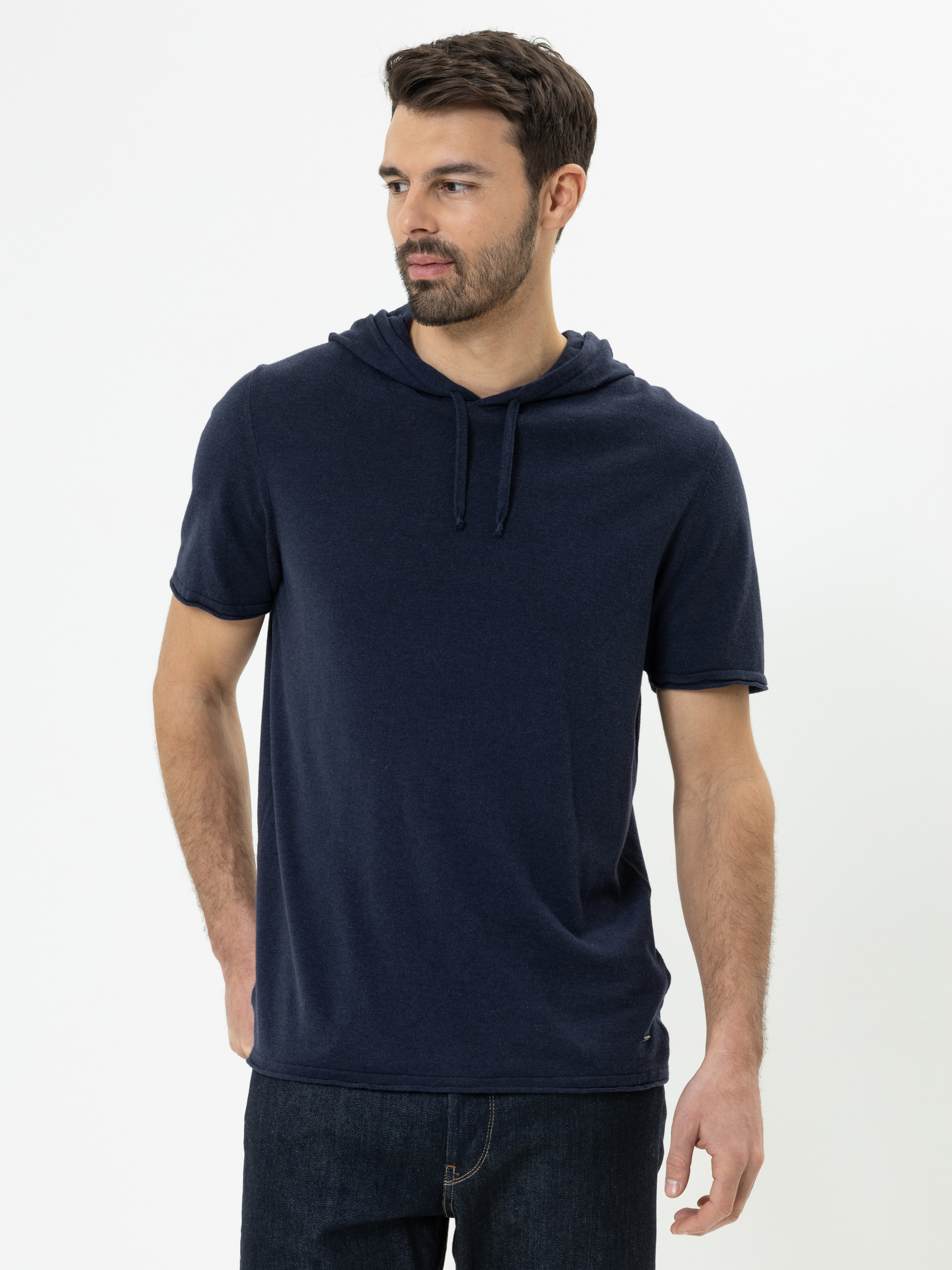 Ein bärtiger Mann, der ein kurzärmeliges, dunkelblaues Kapuzen-T-Shirt und dunkle Jeans trägt, steht vor einem schlichten weißen Hintergrund und schaut mit neutralem Blick leicht zur Seite.