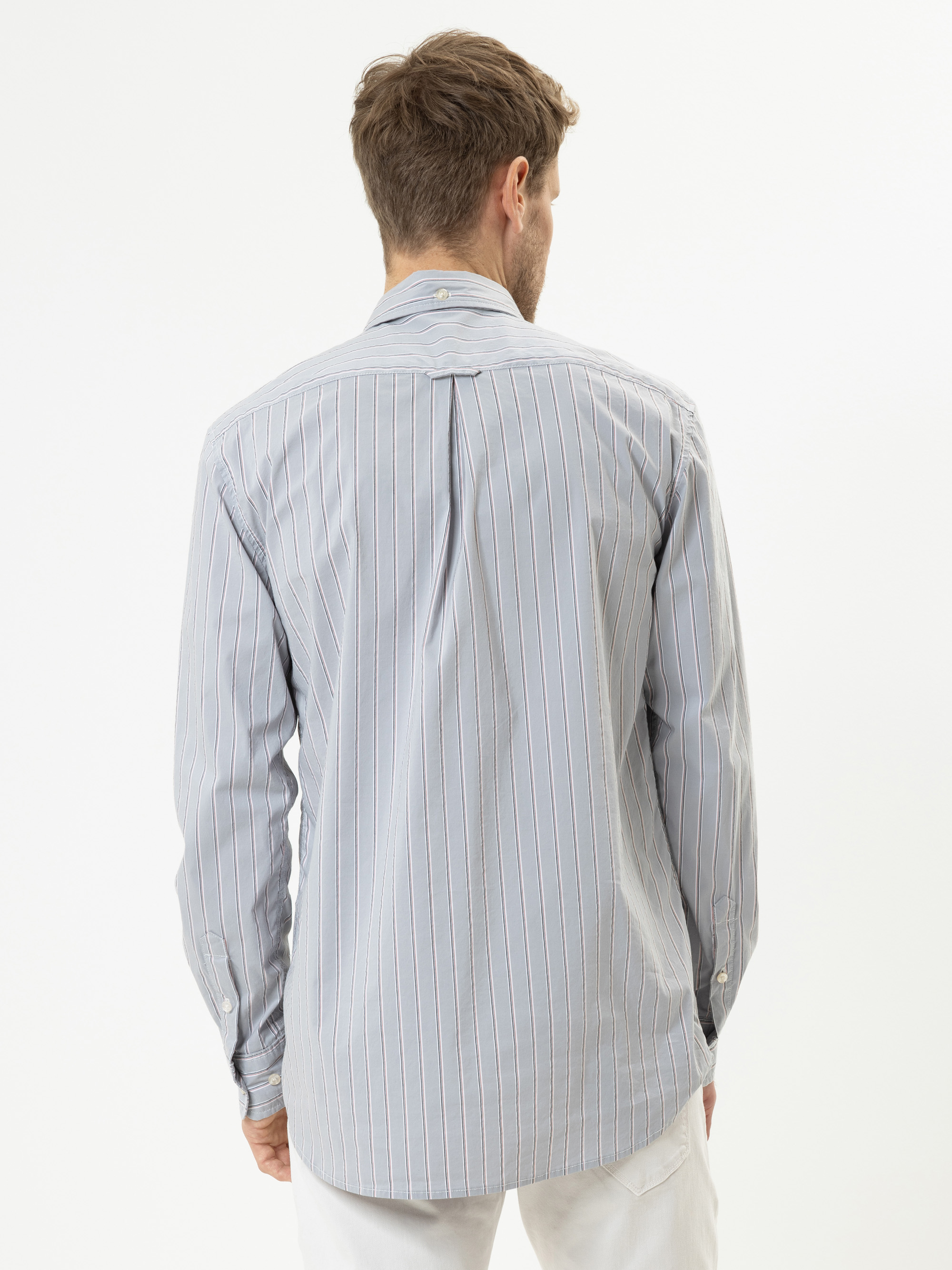 Ein Mann steht mit dem Rücken zur Kamera und trägt ein langärmeliges, hellblau-weißes, senkrecht gestreiftes Hemd und eine weiße Hose vor einem schlichten weißen Hintergrund.