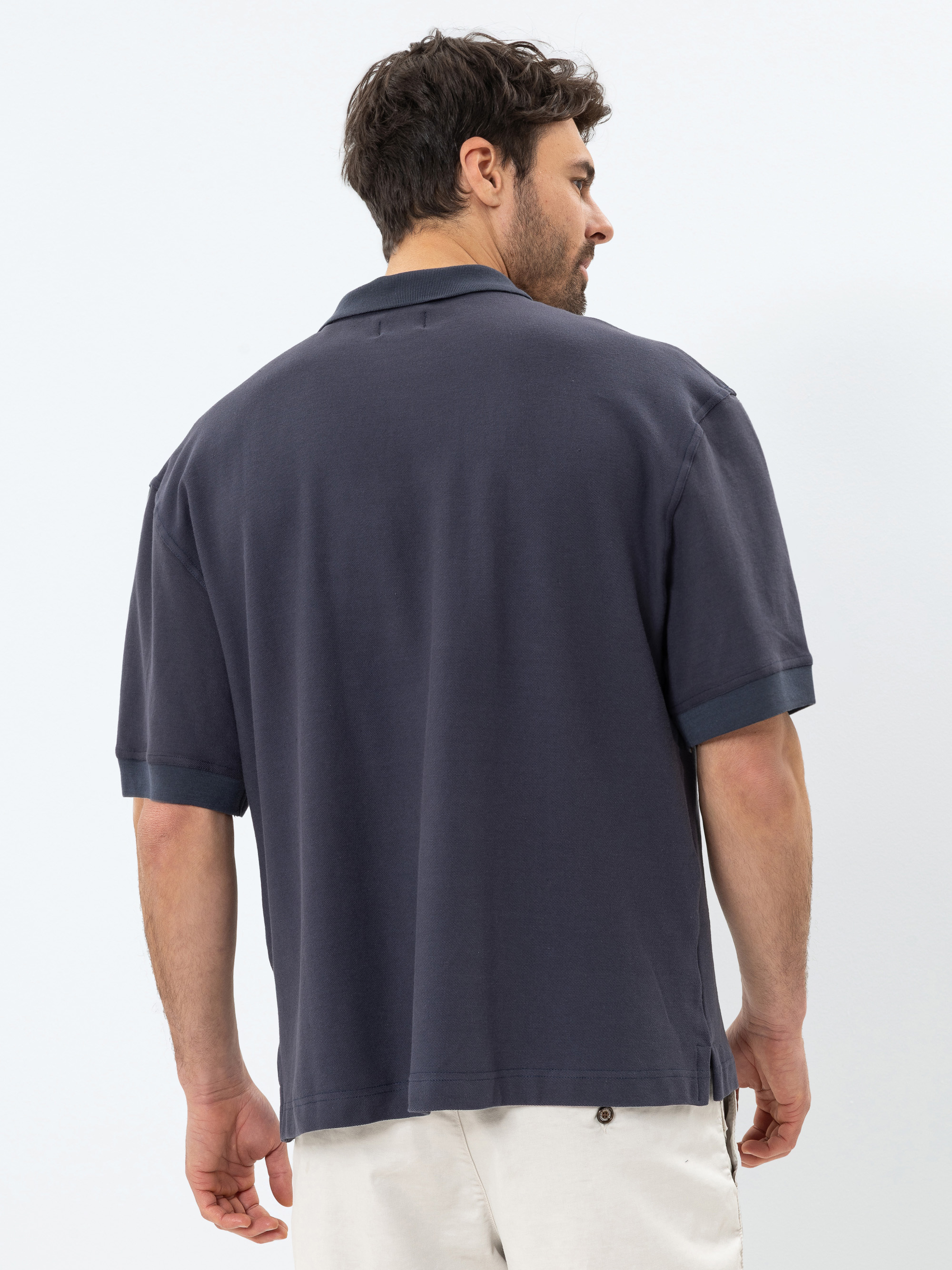 Ein Mann mit kurzen dunklen Haaren wird von hinten gezeigt, der ein locker sitzendes, kurzärmeliges, dunkelblaues Polohemd und helle Shorts trägt und vor einem schlichten weißen Hintergrund steht.