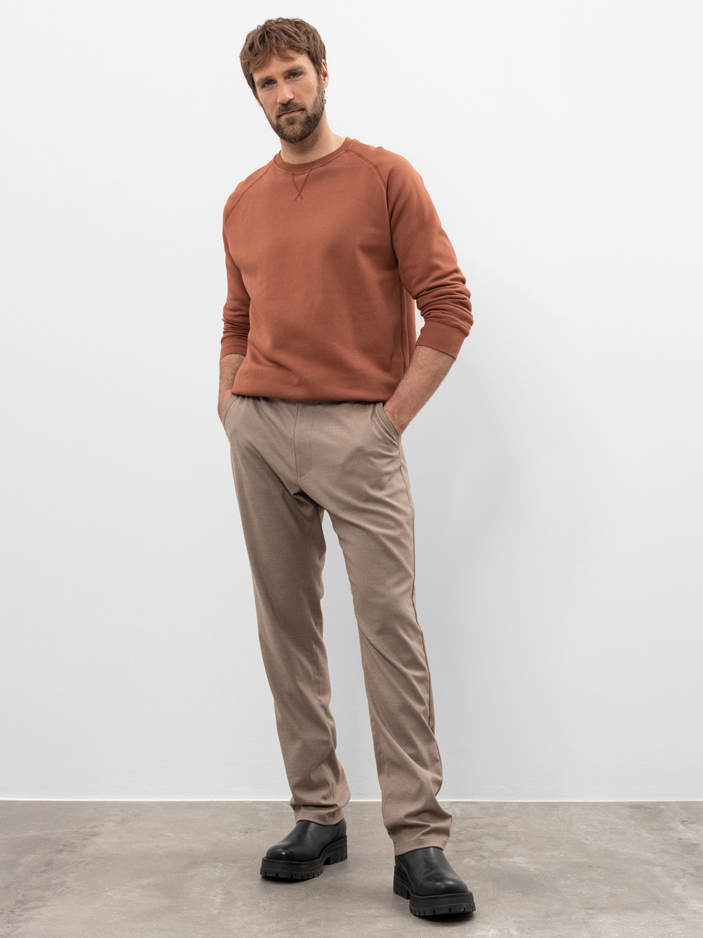 Ein Mann mit hellbraunem Haar und Bart steht vor einer schlichten weißen Wand und trägt ein rostfarbenes Sweatshirt, eine beigefarbene Hose und schwarze Stiefel. Seine Hände stecken in den Taschen und er hat einen neutralen Gesichtsausdruck.