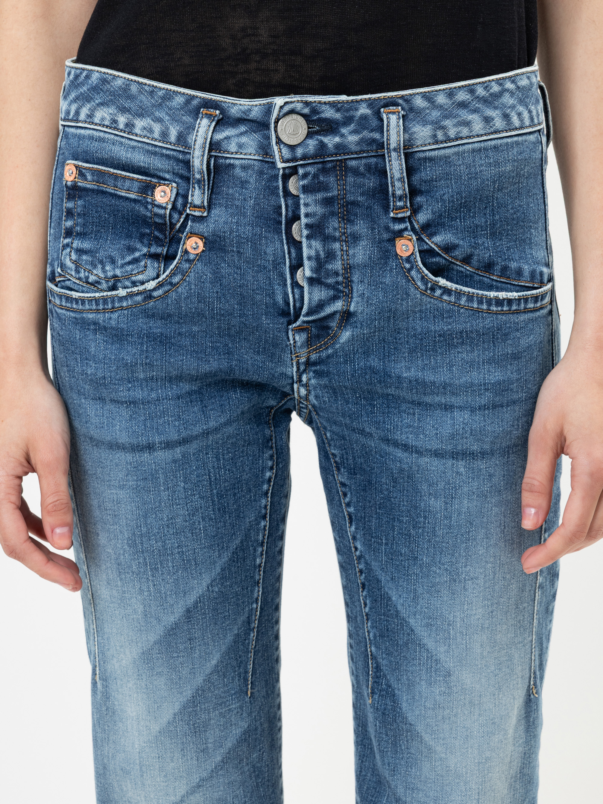 Nahaufnahme einer Person, die eine blaue Jeans und ein schwarzes Oberteil trägt, mit sichtbaren Knöpfen und Nähten. Die Hände der Person liegen entspannt an der Seite.