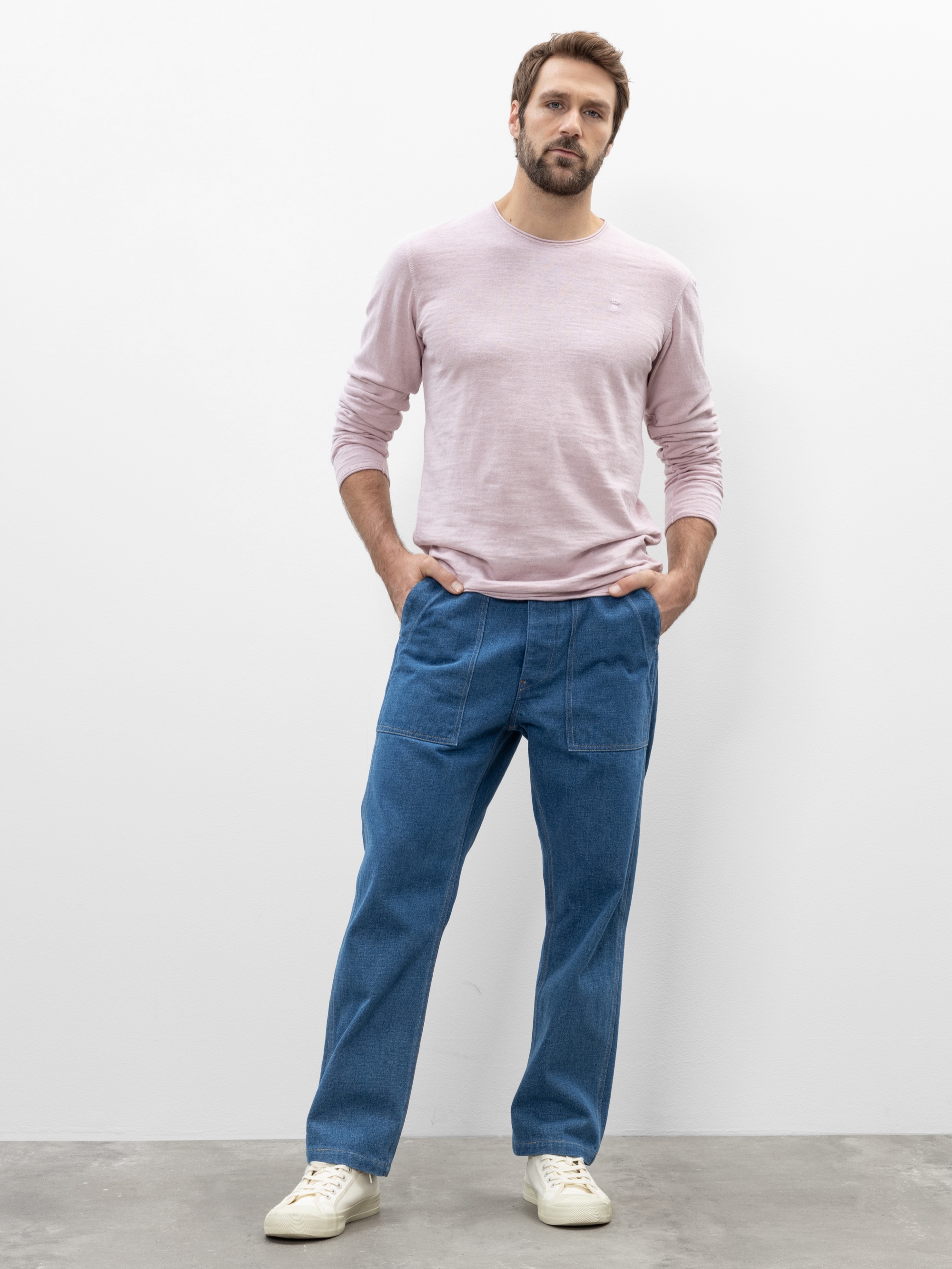 Ein Mann steht vor einer schlichten weißen Wand. Er trägt ein hellrosa, langärmeliges Hemd, eine blaue Jeans mit vorderen Nähten und großen Taschen sowie weiße Turnschuhe. Seine Hände sind in den Taschen und er hat einen neutralen Gesichtsausdruck.