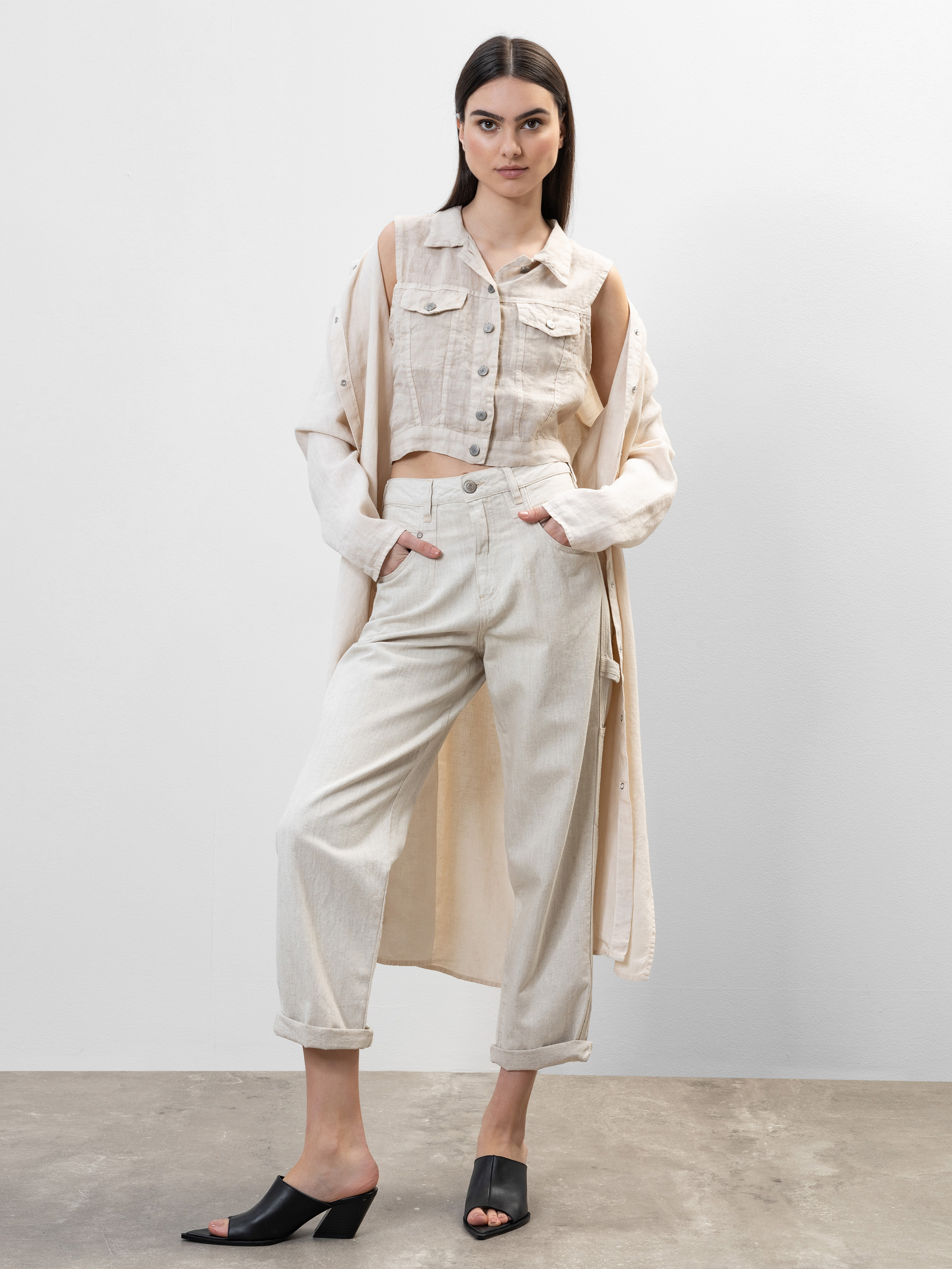 Eine Frau steht selbstbewusst vor einem schlichten, hellen Hintergrund. Sie trägt eine hellbeige ärmellose Kurzjacke, eine dazu passende Hose mit hoher Taille, einen langen beigen Mantel, der über ihre Schultern fällt, und schwarze Pantoletten mit Abs