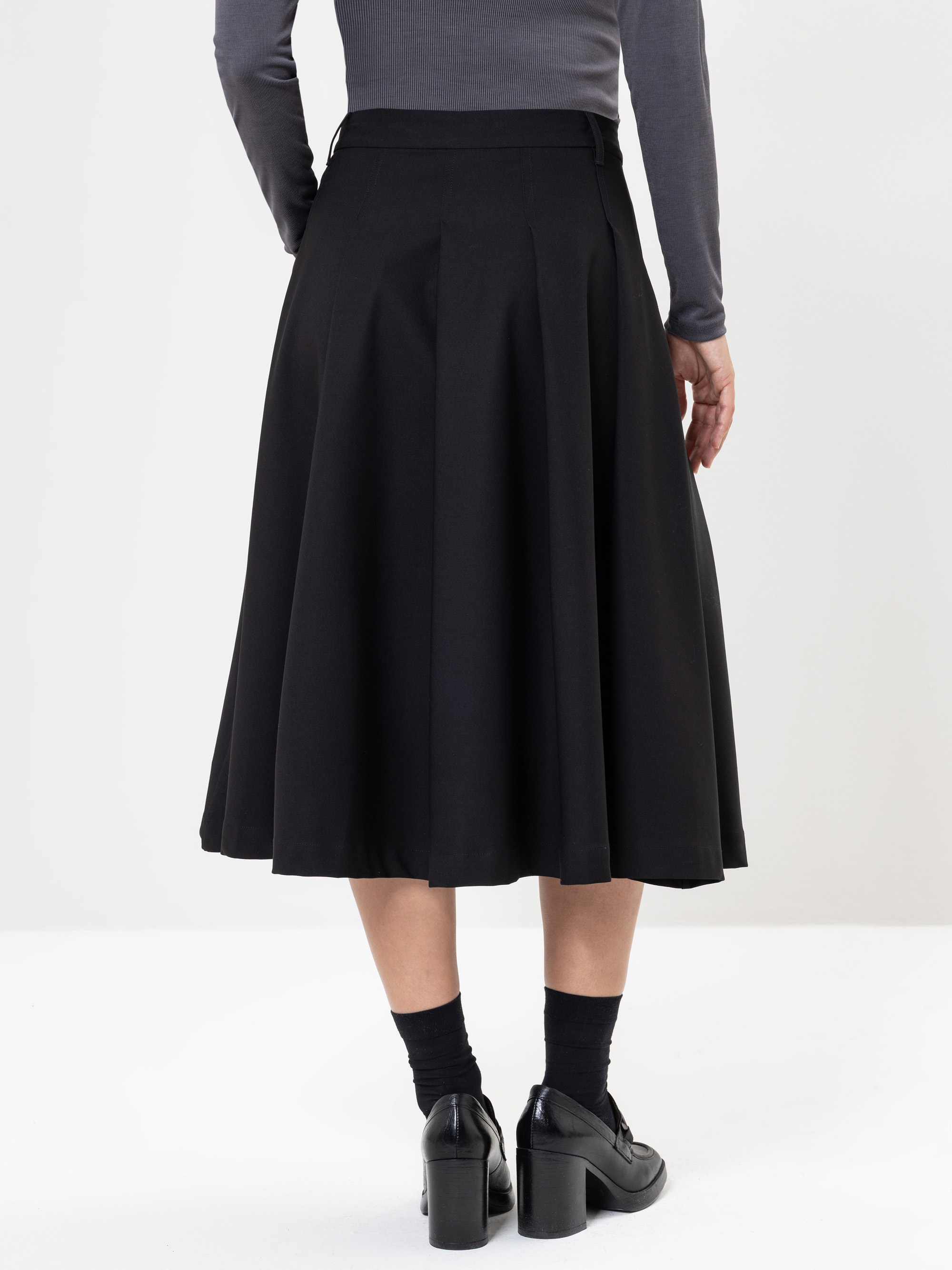 Eine Person, die ein langärmeliges graues Oberteil und einen schwarzen, knielangen Faltenrock trägt, steht mit dem Gesicht zur Seite. Sie trägt schwarze Socken und schwarze Schuhe mit Absätzen vor einem schlichten weißen Hintergrund.