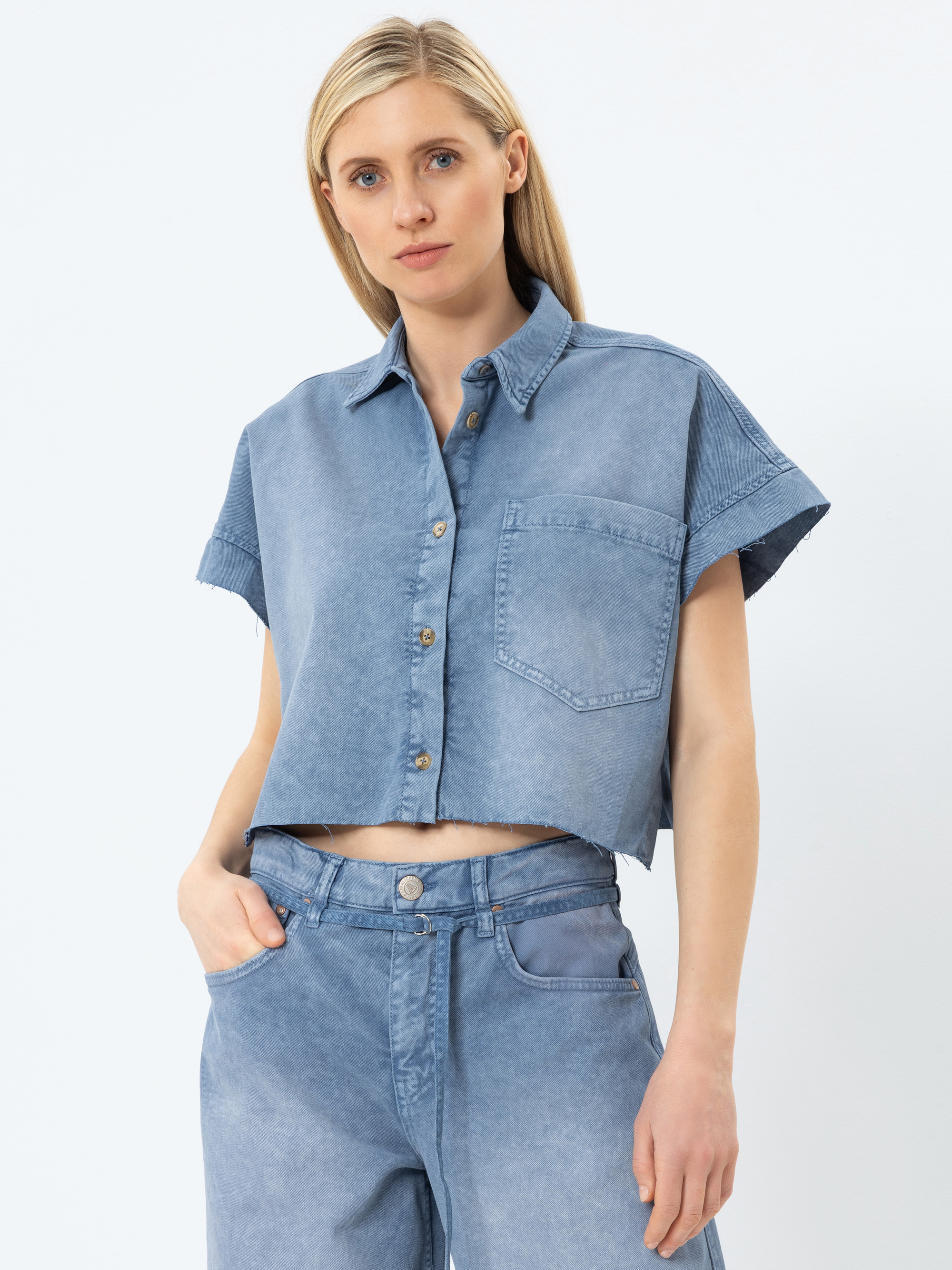 Eine Frau mit glattem blondem Haar trägt ein hellblaues, kurzärmeliges, kurzgeschnittenes Jeanshemd mit Knöpfen und einer Brusttasche, gepaart mit einer hochgeschlossenen, locker sitzenden Bluejeans. Sie hat eine Hand in der Tasche und blickt in die Ka