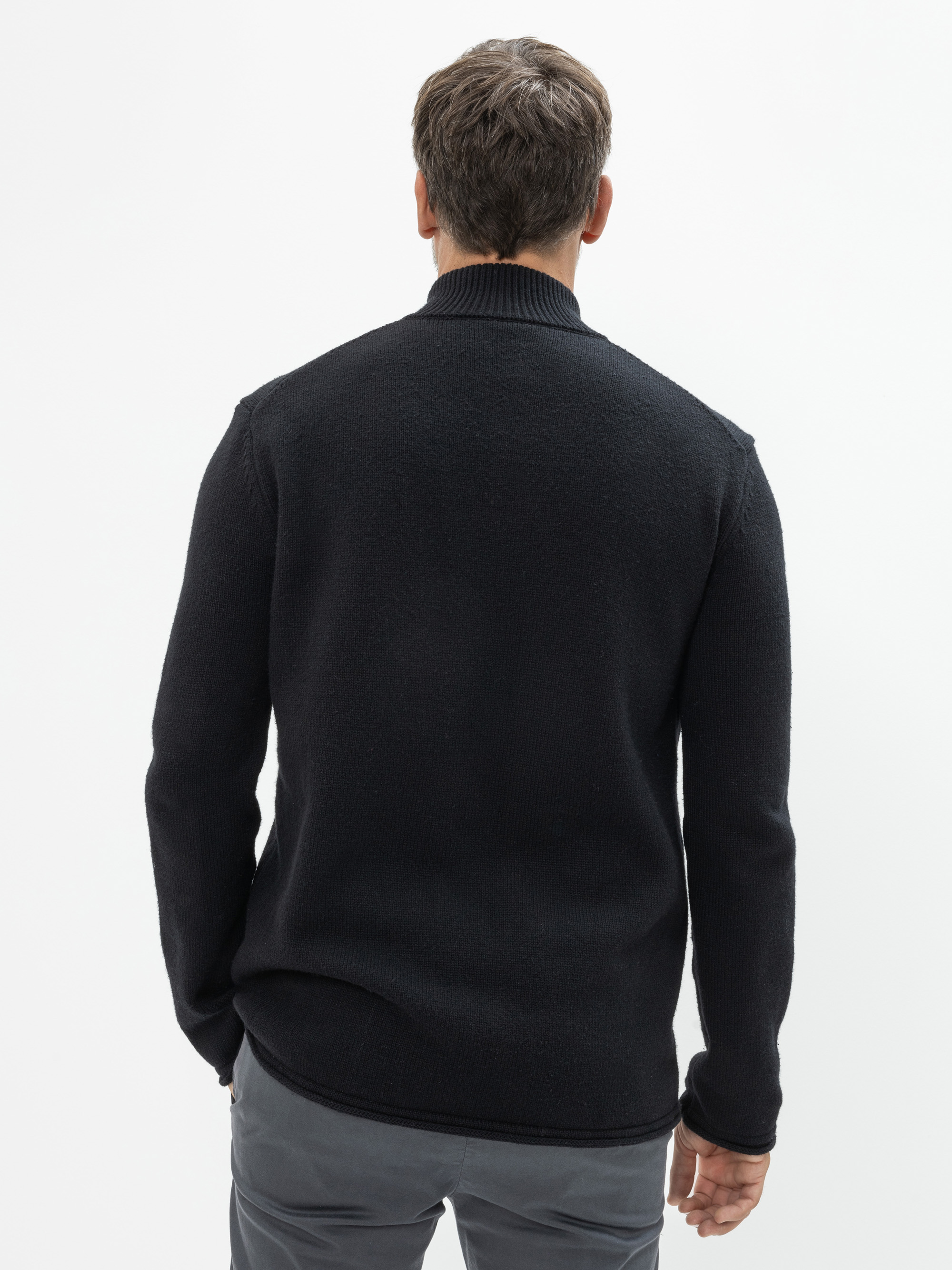 Ein Mann steht von der Kamera abgewandt in einem schwarzen Pullover und einer grauen Hose vor einem schlichten weißen Hintergrund.
