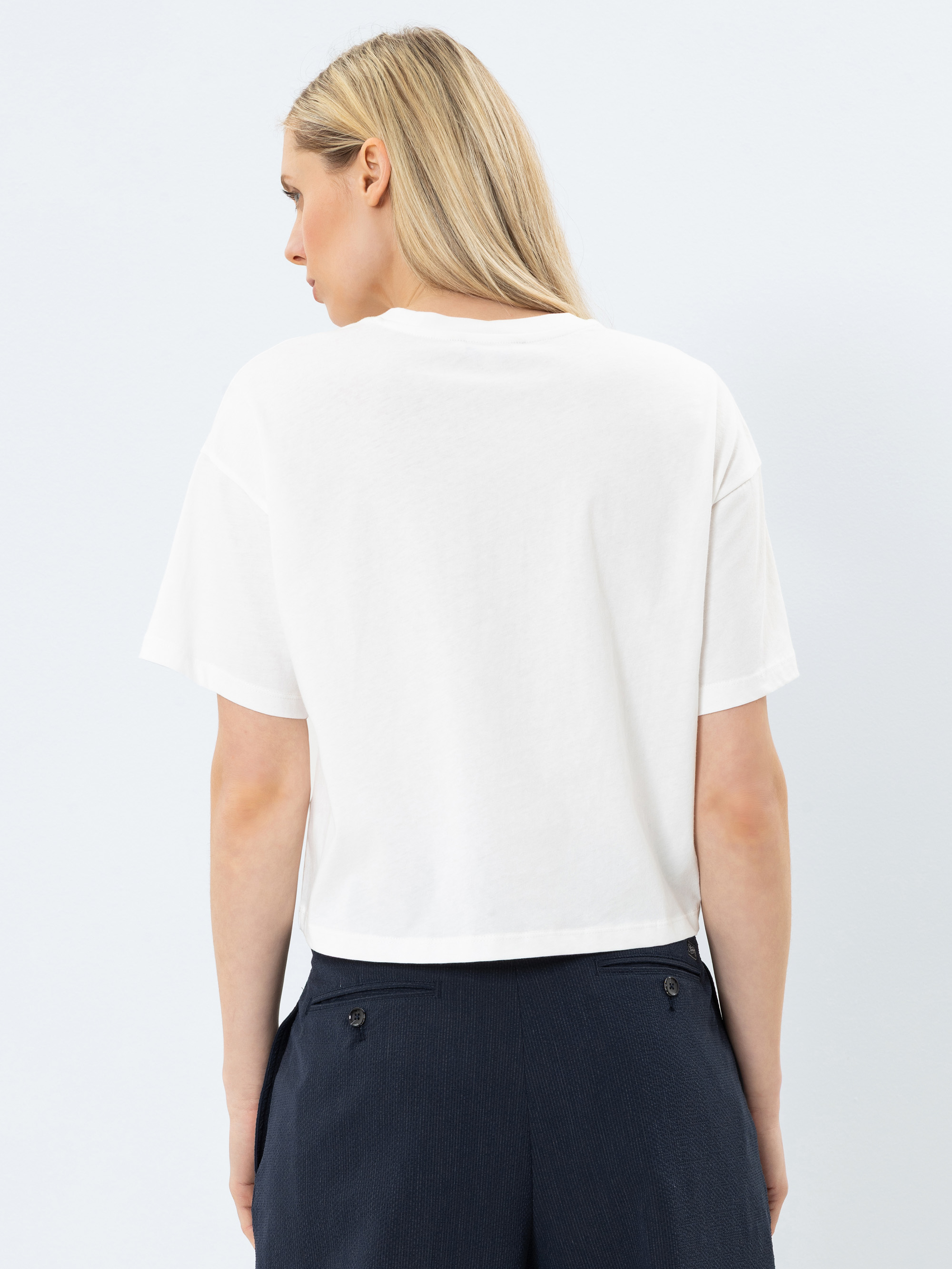Eine Frau mit langen blonden Haaren steht mit dem Rücken zur Kamera und trägt ein schlichtes weißes kurzärmeliges T-Shirt und eine dunkle Hose. Der Hintergrund ist hell und einfarbig.