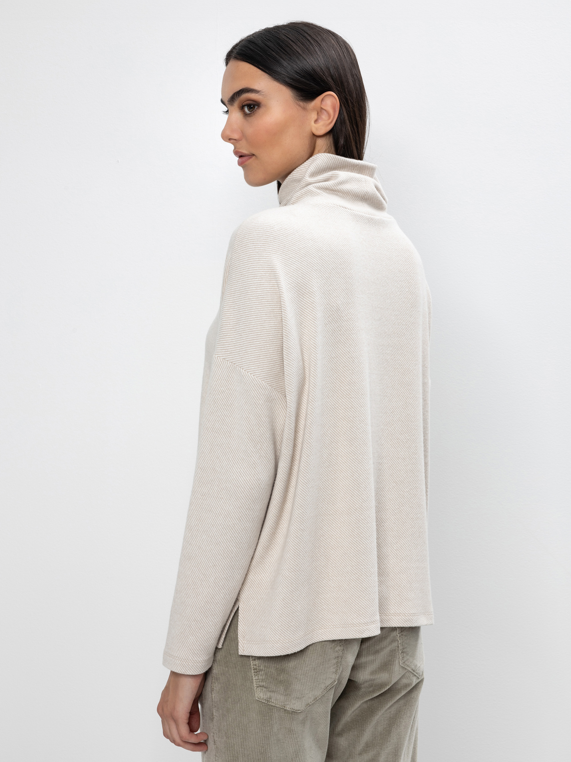 Frau mit glattem dunklem Haar, bekleidet mit einem hellbeigen Rollkragenpullover und einer hellolivfarbenen Hose, die vor einem schlichten weißen Hintergrund steht und über ihre Schulter schaut.