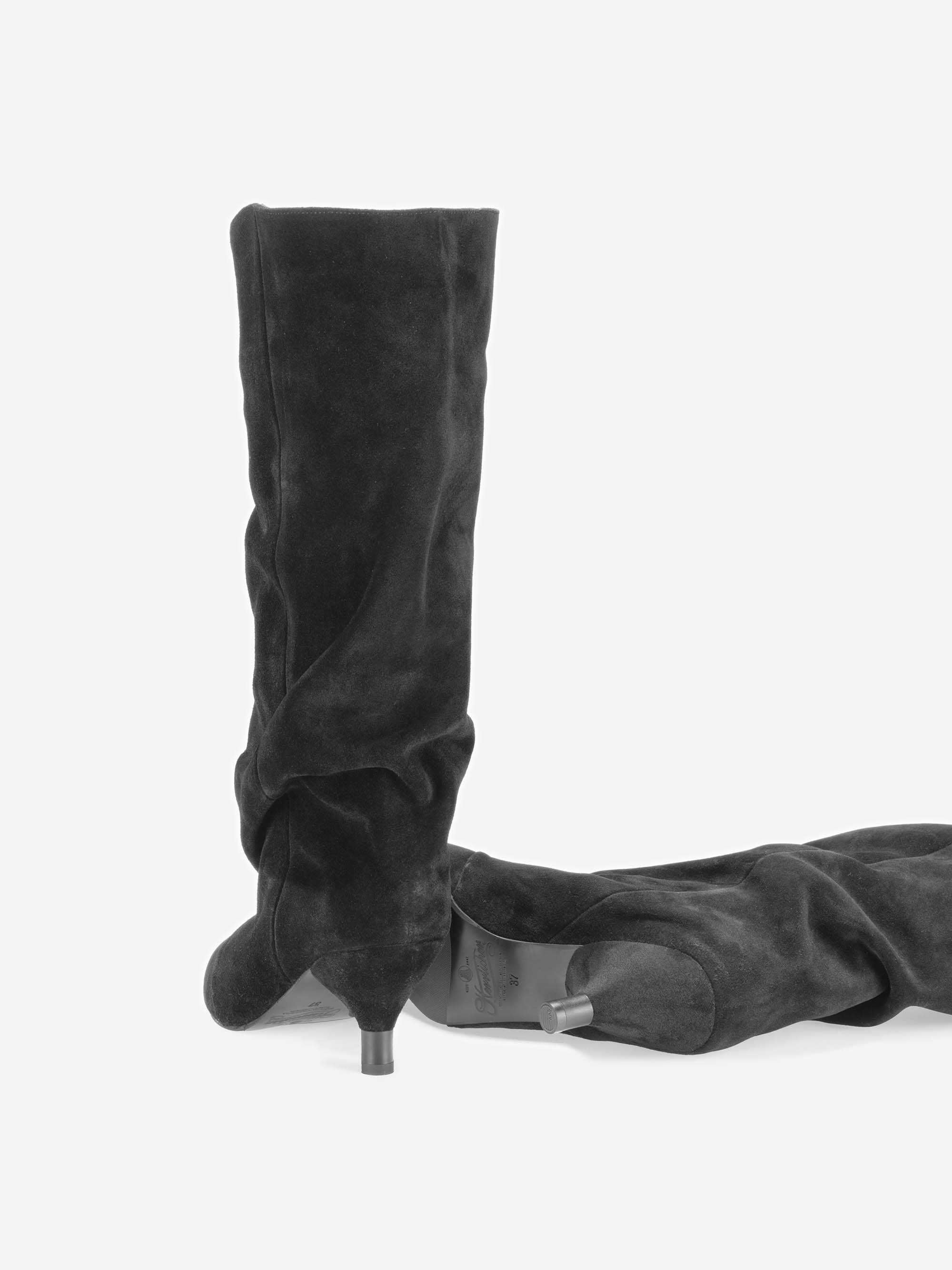 Ein Paar schwarze, kniehohe Wildlederstiefel mit spitzen Zehen und schlanken, mittelhohen Absätzen. Ein Stiefel steht aufrecht, während der andere auf der Seite liegt; beide befinden sich vor einem schlichten weißen Hintergrund.