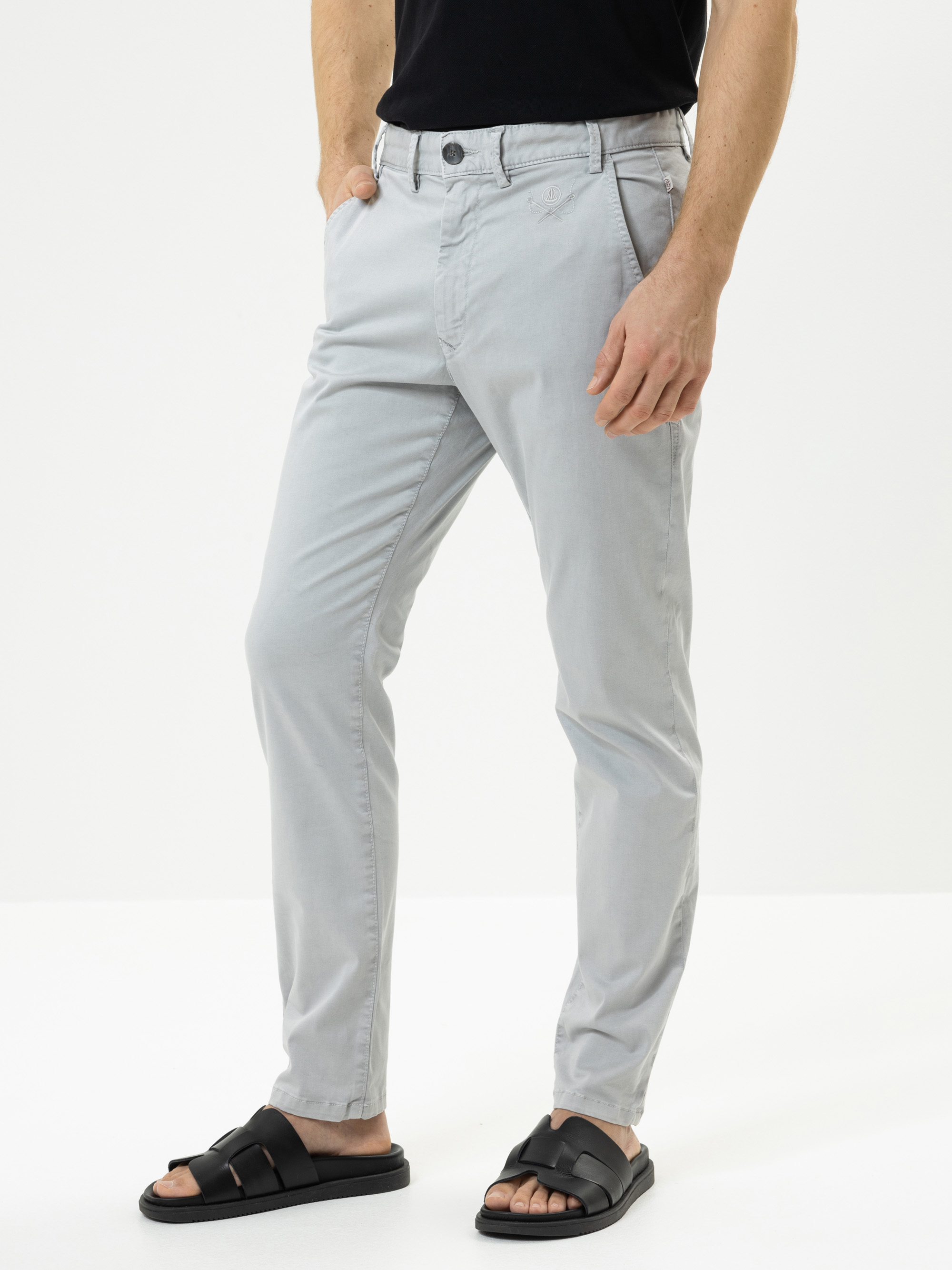 Eine Person, die eine hellgraue Chinohose, ein schwarzes T-Shirt und schwarze Sandalen trägt, steht mit den Händen in den Taschen vor einem schlichten weißen Hintergrund.