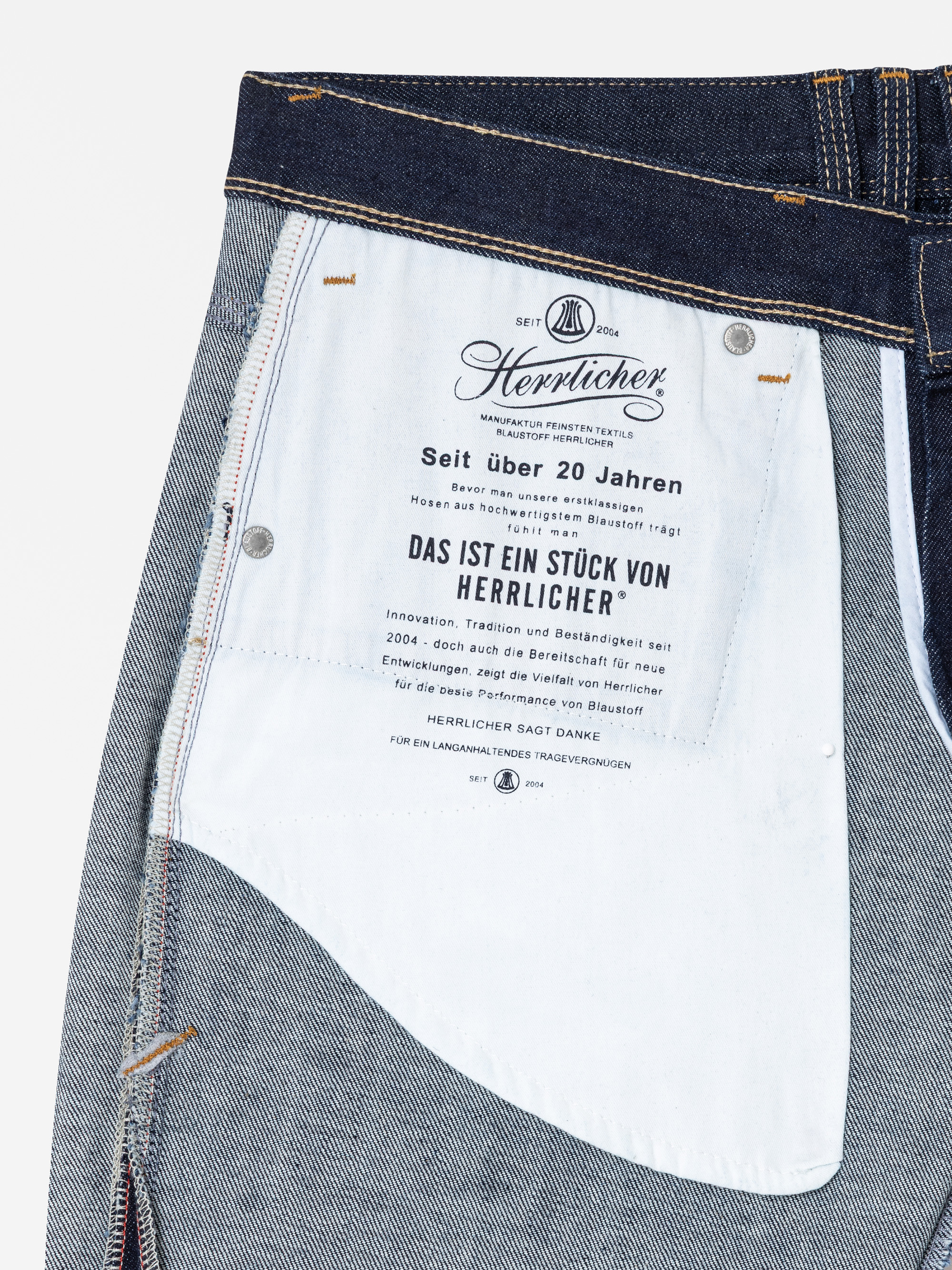 Nahaufnahme der Innentasche einer Jeans mit weißem Taschenfutter und schwarzem Aufdruck des deutschen Textes und des Herrlicher-Markenzeichens sowie sichtbaren Nähten und Denim-Stoff.
