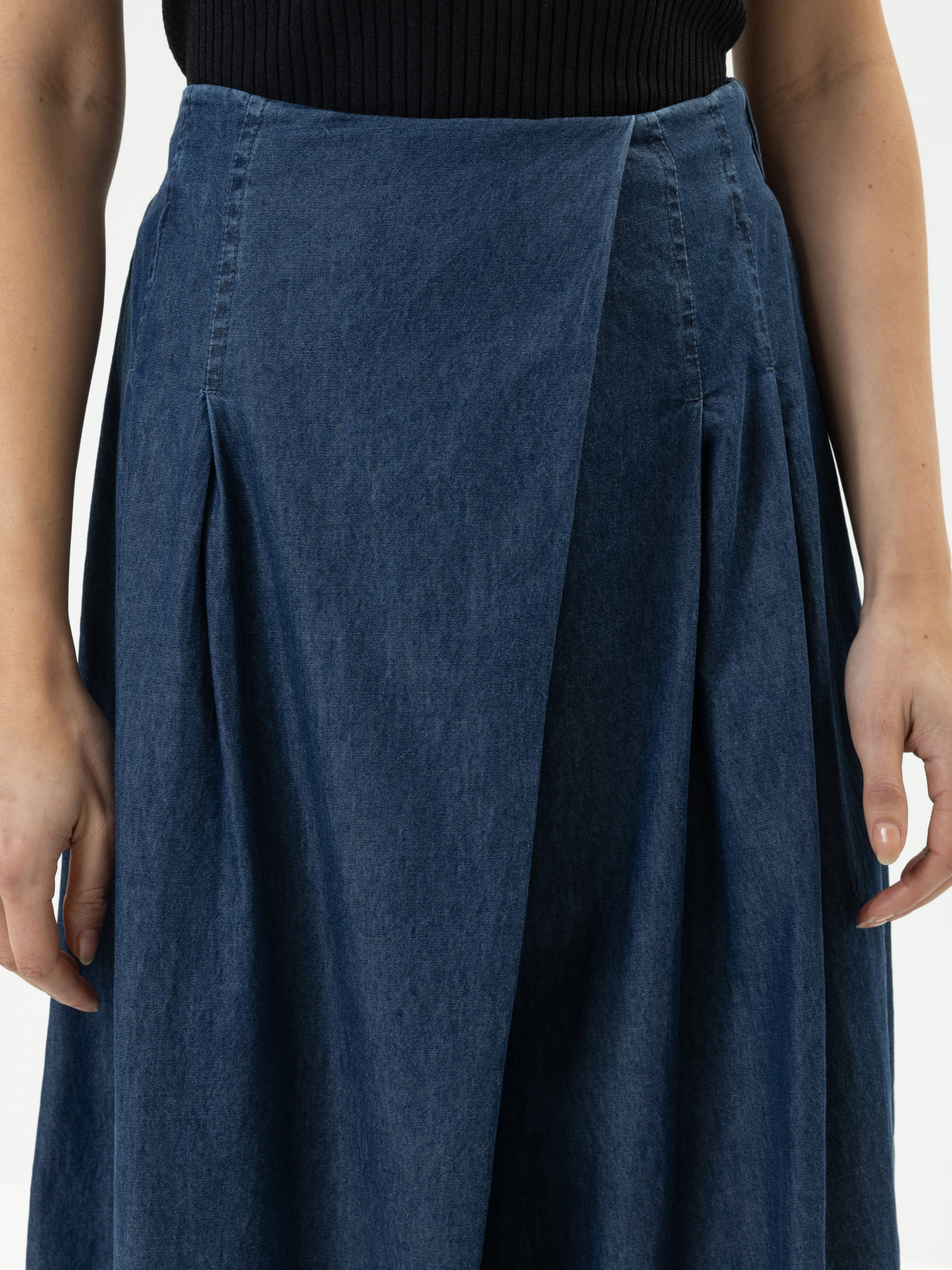 Nahaufnahme einer Person, die einen dunkelblauen Jeans-Faltenrock und ein schwarzes geripptes Oberteil trägt. Die Arme der Person liegen entspannt an der Seite, nur der untere Teil des Oberkörpers und die Hände sind zu sehen.