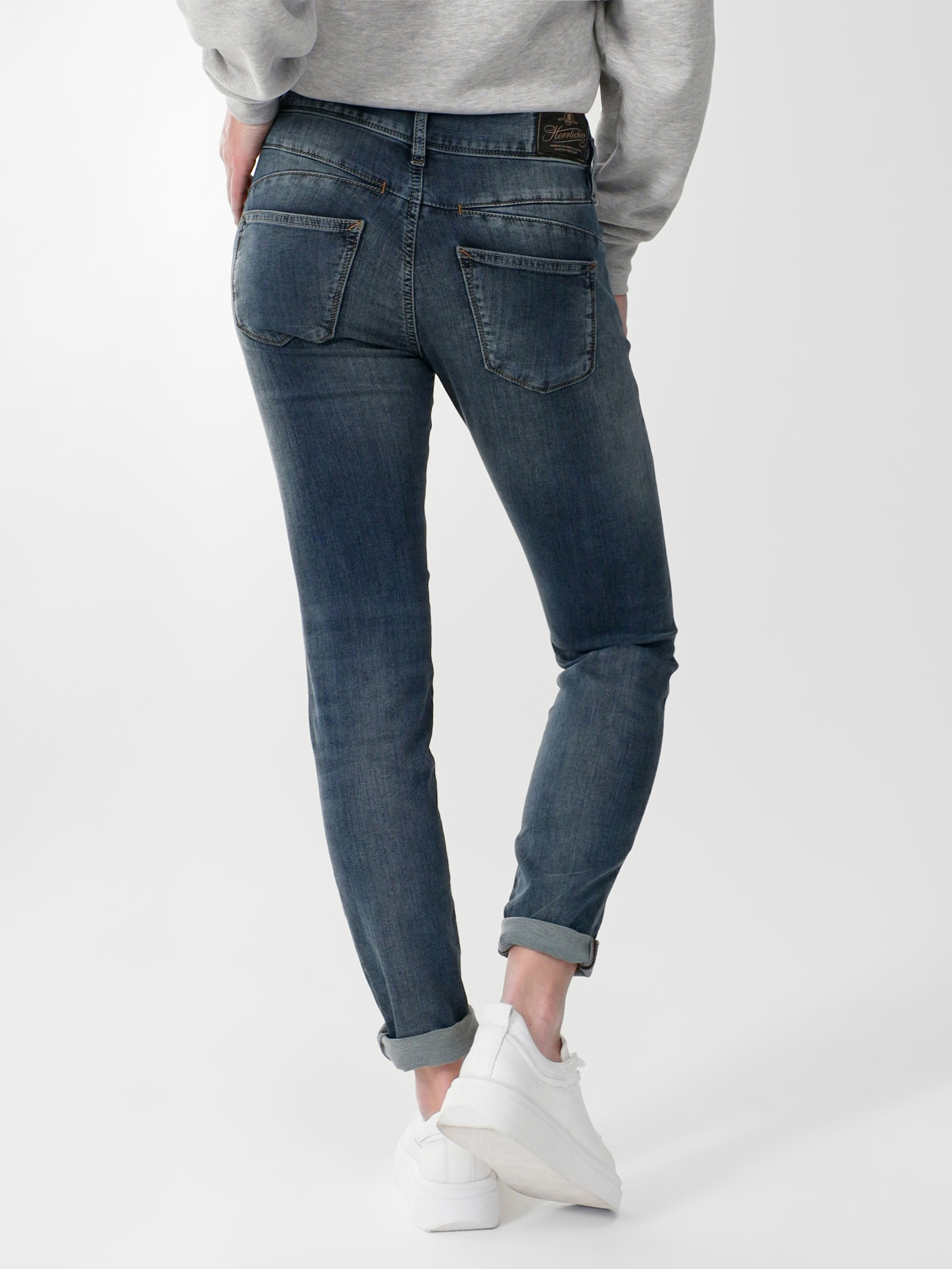 Eine Person, die blaue Jeans mit Bündchen, ein graues Sweatshirt und weiße Turnschuhe trägt, steht mit dem Rücken zur Kamera vor einem schlichten weißen Hintergrund.