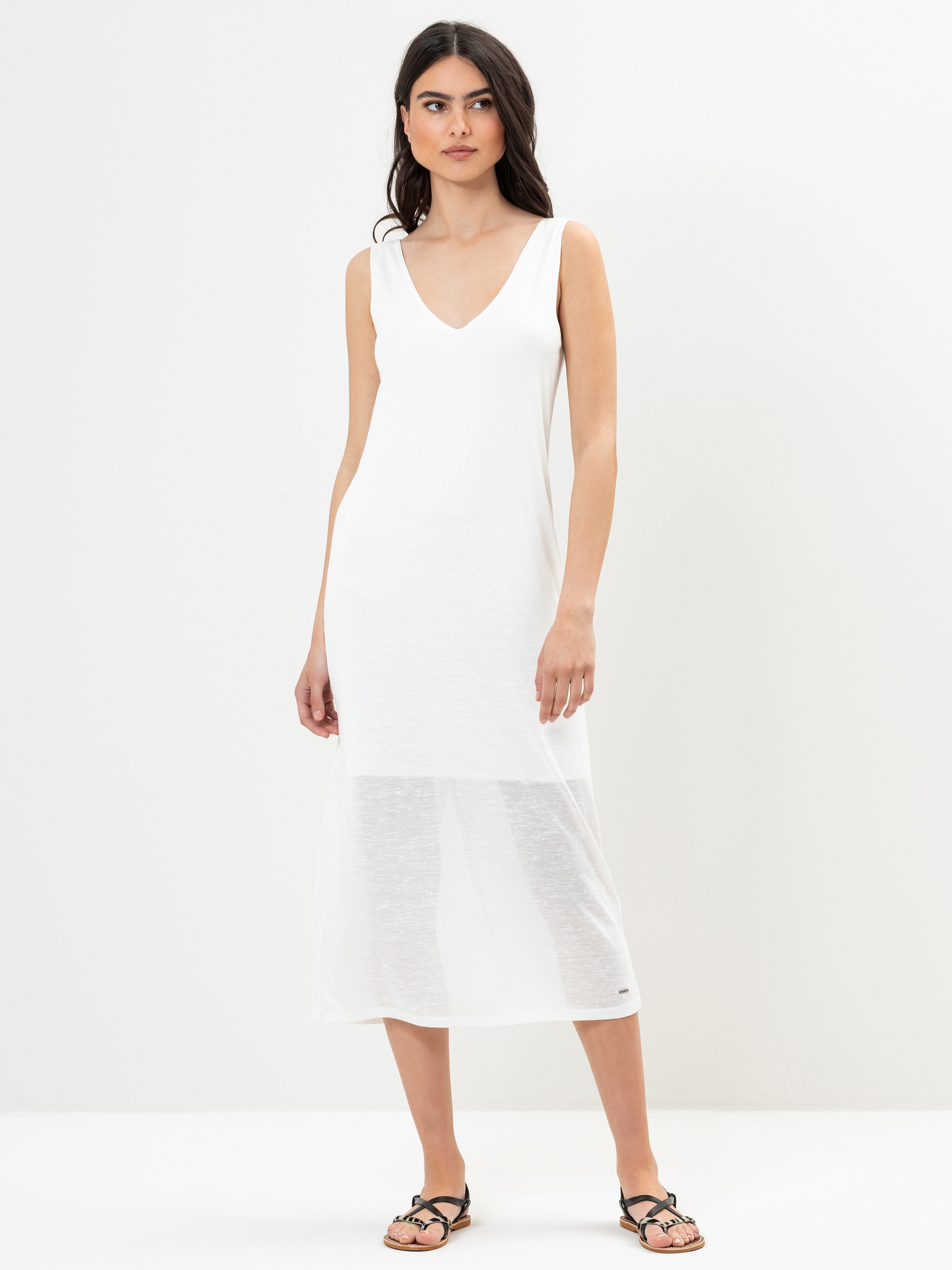 Eine Frau steht vor einem schlichten weißen Hintergrund und trägt ein ärmelloses, weißes, knöchellanges Kleid mit V-Ausschnitt und durchscheinendem Stoff am Saum sowie schwarze Sandalen. Sie hat langes dunkles Haar und einen neutralen Gesichtsausdruc