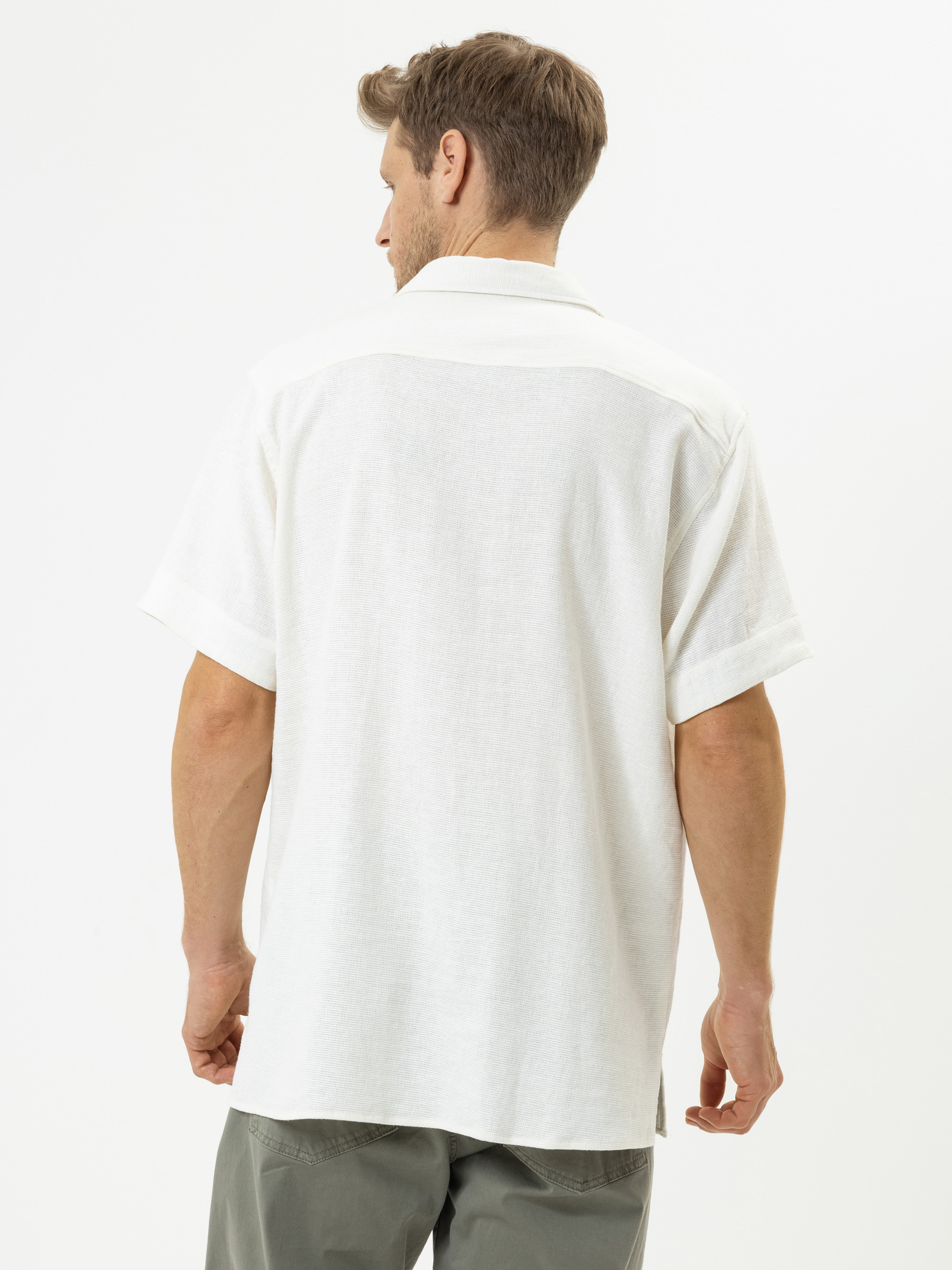 Ein Mann mit hellbraunem Haar, der ein kurzärmeliges weißes Hemd und eine graugrüne Hose trägt, steht mit dem Gesicht zur Kamera vor einem schlichten weißen Hintergrund.