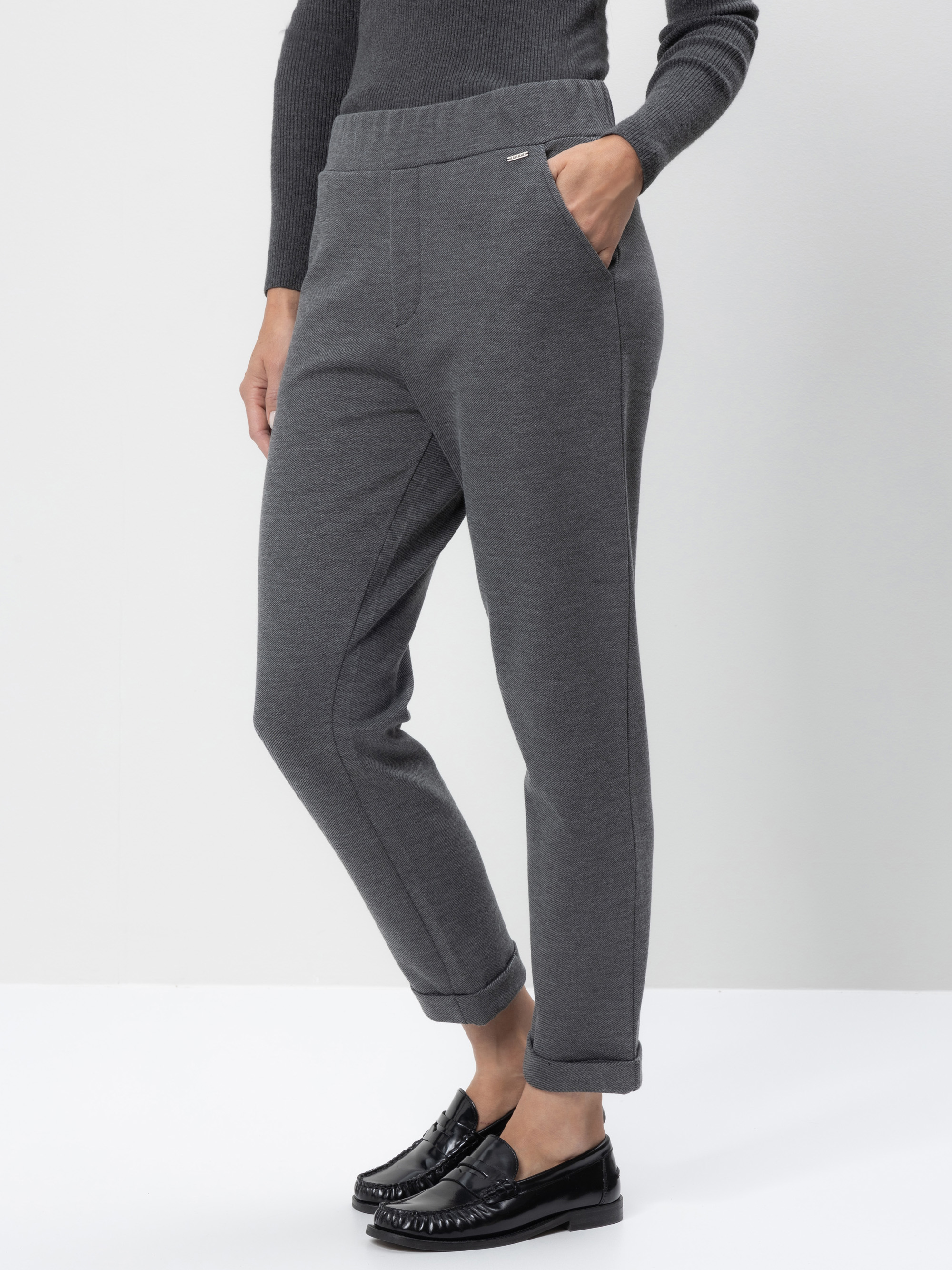 Eine Person, die eine dunkelgraue, knöchellange Jogginghose mit Seitentaschen, einen gerippten dunklen Pullover und schwarze Slipper trägt, steht vor einem schlichten weißen Hintergrund.