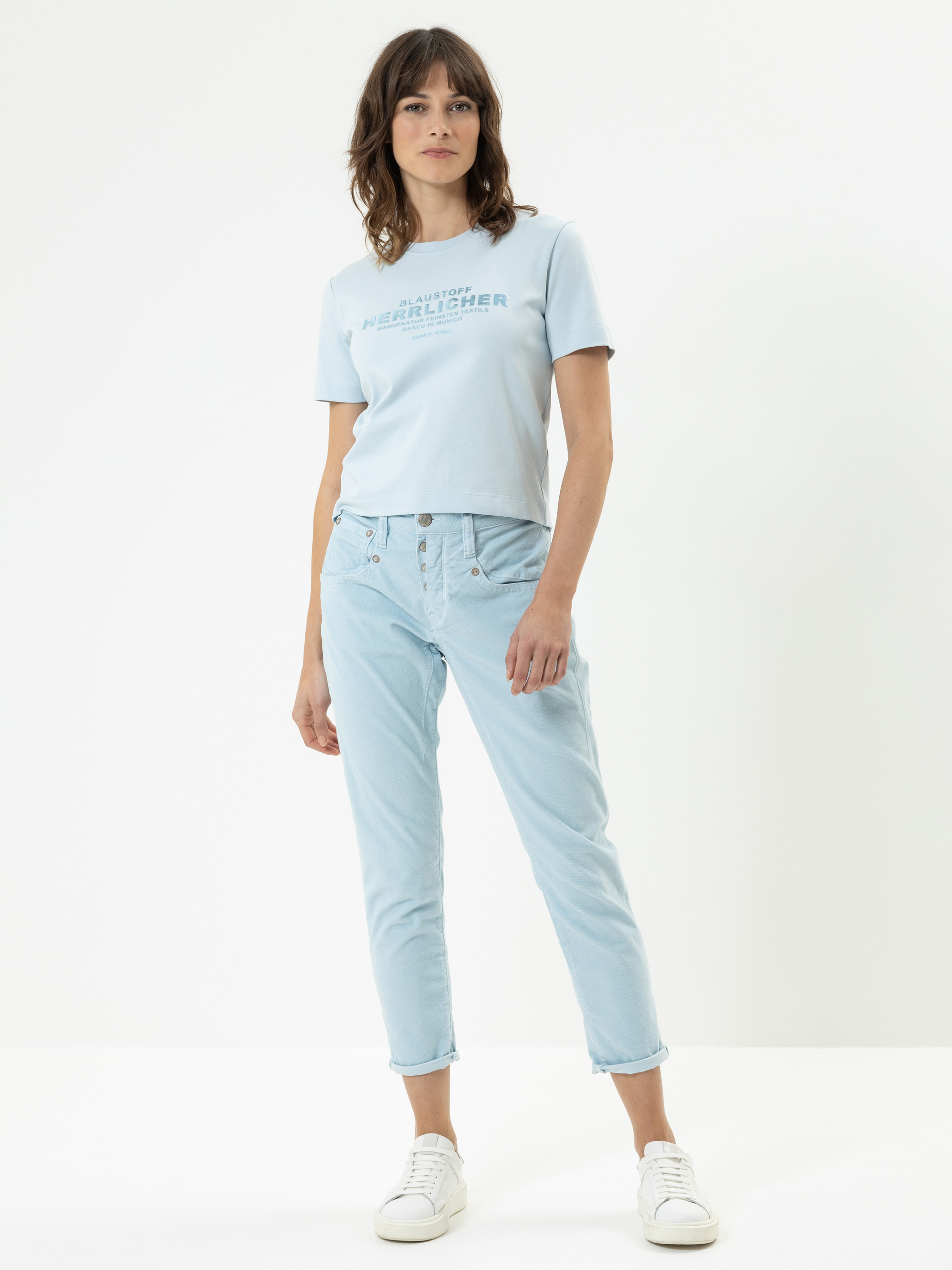 Eine Frau steht vor einem schlichten weißen Hintergrund und trägt ein hellblaues T-Shirt mit Text, hellblaue Jeans und weiße Turnschuhe. Sie hat schulterlanges braunes Haar und eine entspannte Körperhaltung mit einer Hand in der Tasche.