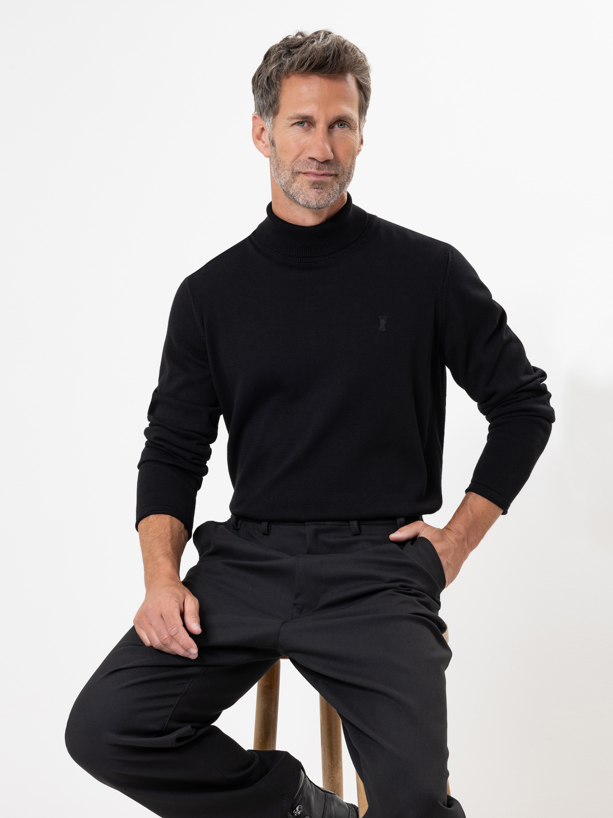 Ein Mann mit kurzen braunen Haaren und Bart sitzt auf einem Holzschemel vor weißem Hintergrund und trägt einen schwarzen Rollkragenpullover und eine schwarze Hose. Eine Hand ruht auf seinem Knie, und er schaut direkt in die Kamera.