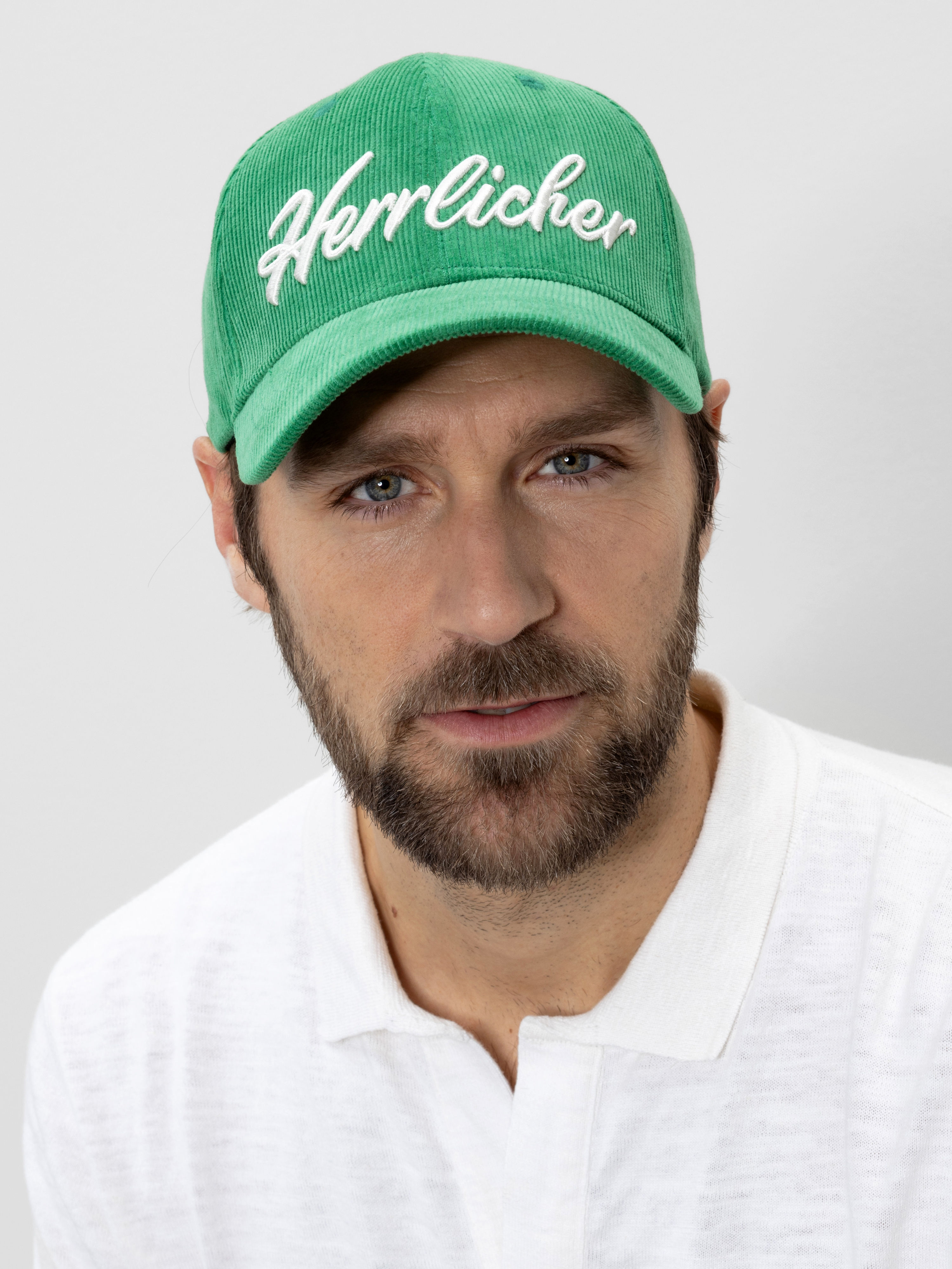 Ein Mann mit Bart und Schnurrbart trägt eine grüne Mütze, auf der das Wort Herrlicher weiß aufgestickt ist, und ein weißes Hemd mit Kragen. Er blickt direkt in die Kamera vor einem unifarbenen Hintergrund.