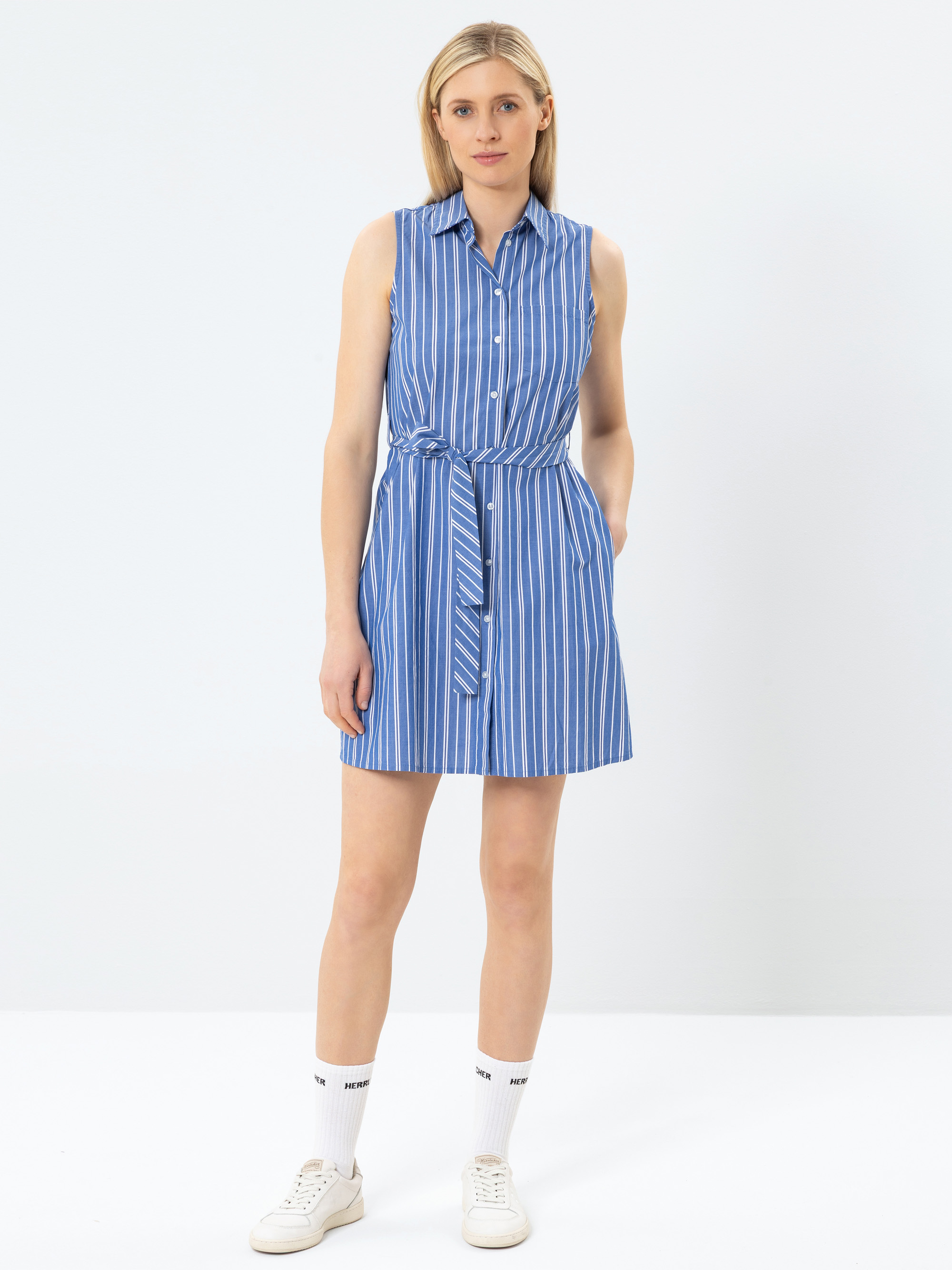 Eine Frau steht vor einem einfarbigen Hintergrund und trägt ein ärmelloses, blau-weißes, vertikal gestreiftes Hemdkleid mit einem passenden Bindegürtel, weiße Turnschuhe und weiße Socken. Sie hat helles Haar und einen neutralen Gesichtsausdruck.